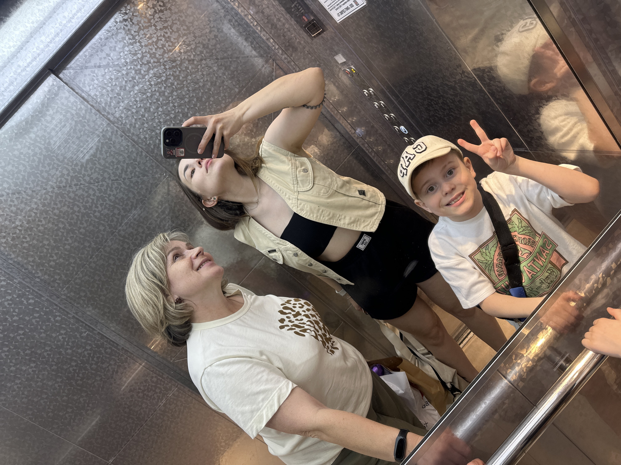 Three individuals are in an elevator taking a photo, likely a selfie, using a cell phone. One person is a woman with blonde hair wearing a white t-shirt with a brown animal print. Another is a young woman with long dark hair wearing a black top, black shorts, and a light-colored jacket. The third person is a young boy wearing a white baseball cap, a white t-shirt with a design, and a shoulder bag, holding up two fingers in a peace sign. They are inside an elevator with patterned metal walls and a mirrored panel. There is evidence of button panel indicating stops for the elevator. The location is indicated to be Perm, Russia.