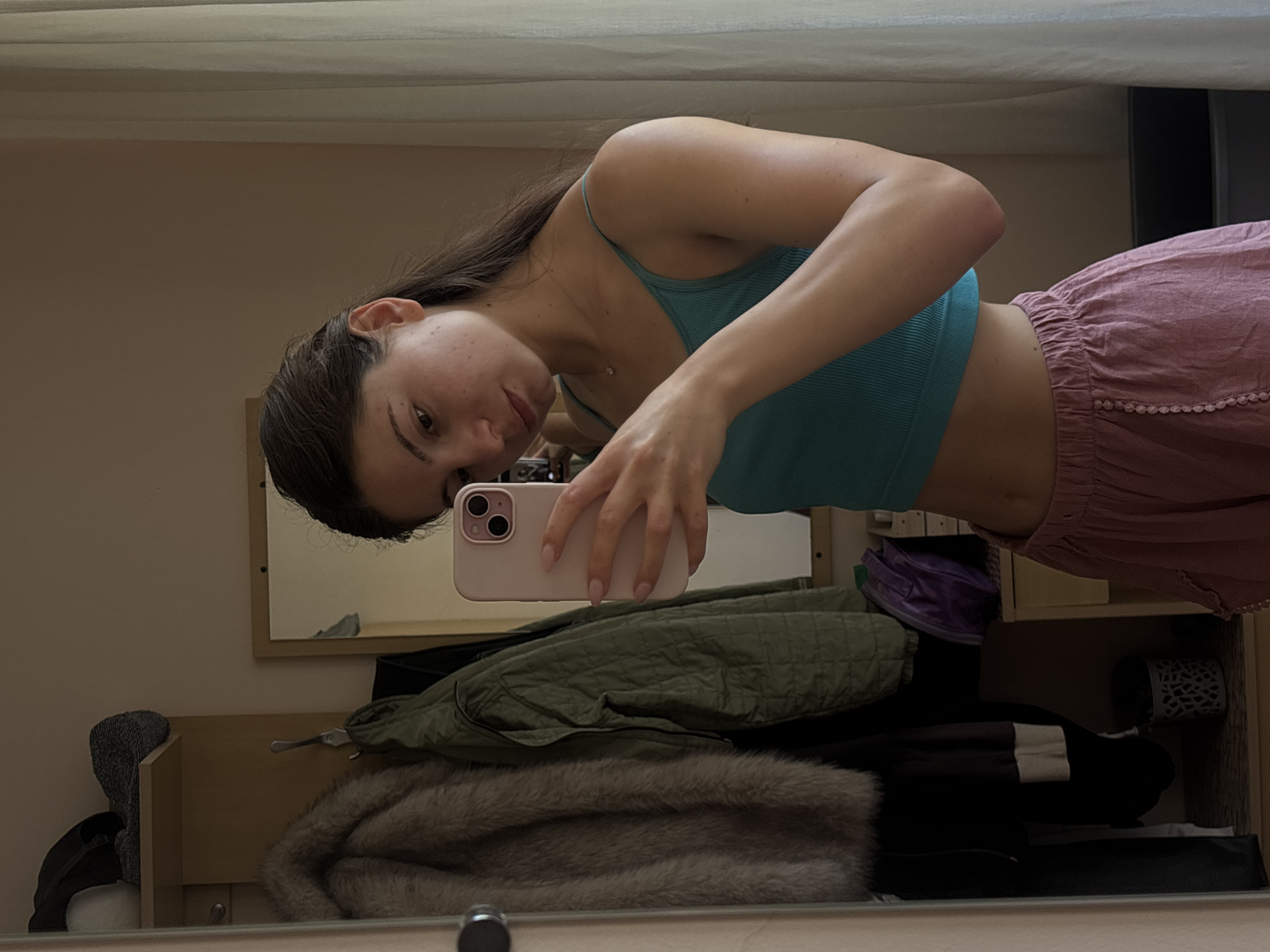 A young woman with long, dark hair pulled back in a ponytail takes a selfie in a mirror. She is wearing a teal ribbed crop top and loose, pink, high-waisted shorts with decorative buttons. She has a delicate necklace on. Her left arm is raised, holding a light pink smartphone.
The background shows a closet or dressing area. To the left of the woman, a fur-lined coat and a green quilted jacket hang on hooks. A purple bag is visible on a shelf above a small dresser. A mirror reflects more of the room, including a window with sheer white curtains on the right. The overall lighting suggests daytime, likely indoors. The environment appears to be a residential setting, possibly a bedroom or dressing room. There are no specific landmarks or indications that place the scene in Varna, Bulgaria.