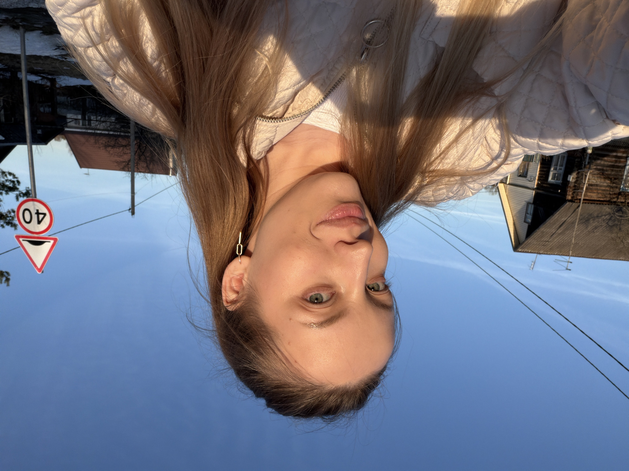The image is an upward-angled selfie of a fair-skinned woman with long, light brown hair wearing a white quilted jacket. The background shows a clear blue sky, power lines, and parts of buildings, including a house with a dark roof and another building with a red roof. A triangular road sign with a "40" speed limit is visible. The photo appears to have been taken outdoors in a residential area, likely in the city of Reshetnikovo, Russia. The woman is the primary subject, with the surrounding environment acting as context.