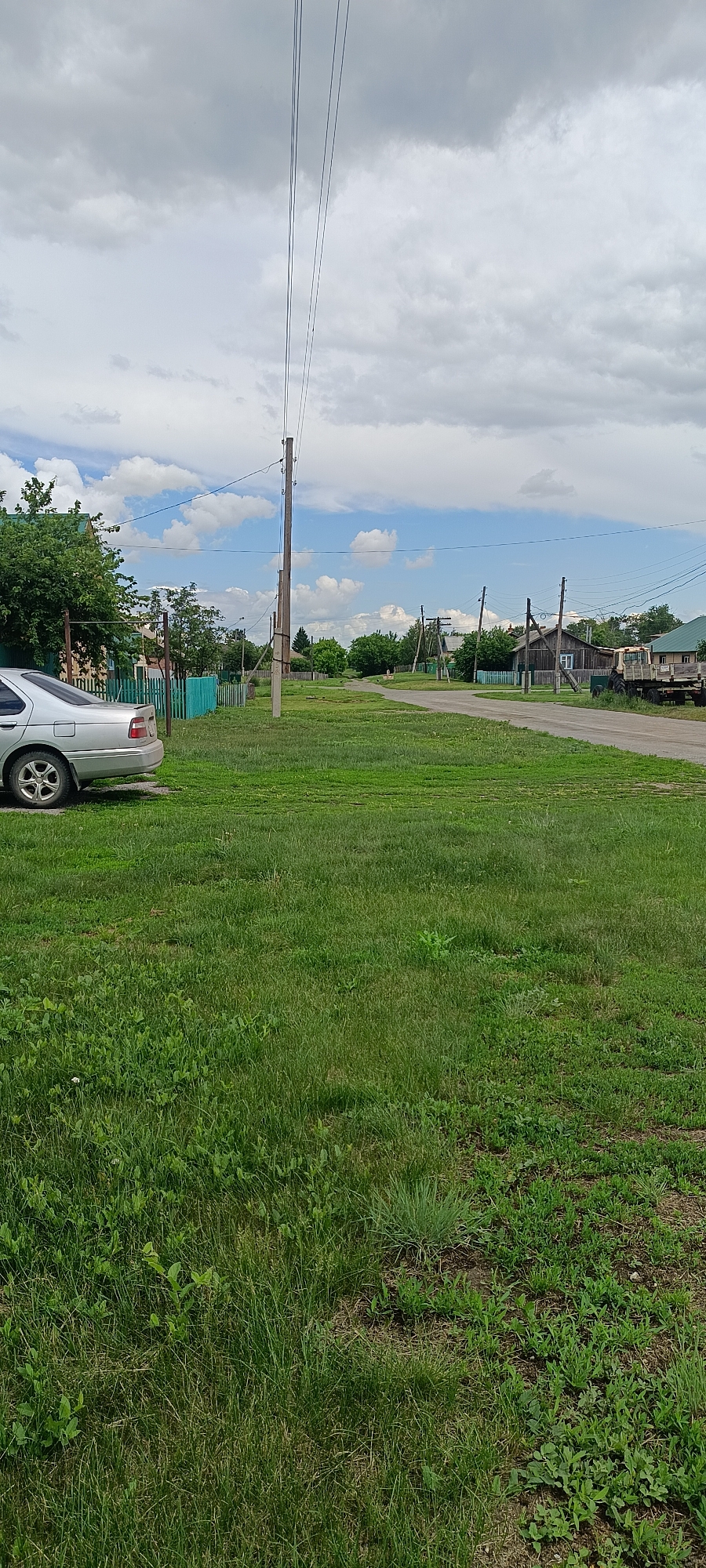 The image depicts a street scene in the village of Kamennoye, Russia, on a cloudy day. A silver sedan is parked on the left side of the frame, next to a grassy area with a teal-colored fence. The street is unpaved and curves to the right, leading into the distance where more houses and trees are visible. Power lines run overhead, supported by wooden poles. A tractor and trailer are parked on the right side of the road. The overall impression is one of a quiet rural setting.