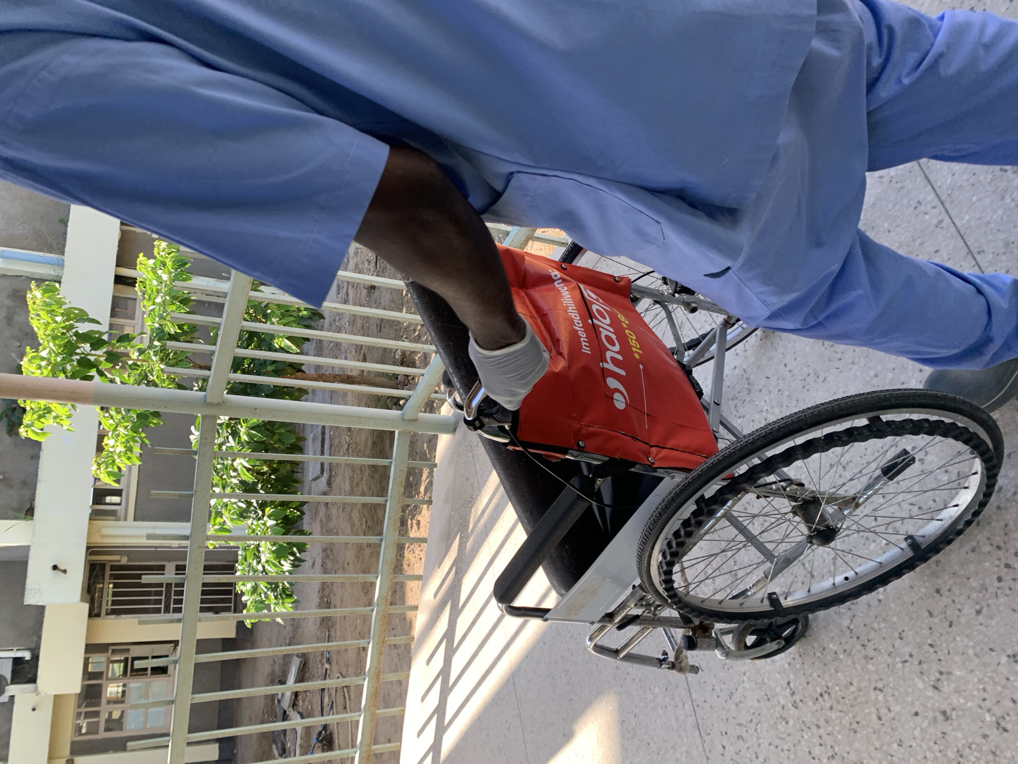 A high-angle shot captures a person in light blue scrubs standing next to a wheelchair. The person's gloved hand rests on the wheelchair's handle. An orange bag with "Halot Medical" printed on it is attached to the back of the wheelchair. The wheelchair's wheel is visible in the foreground, and a railing is partially visible to the left. In the background, there is a building with trees in front of it. The scene is in Dar es Salaam, Tanzania.