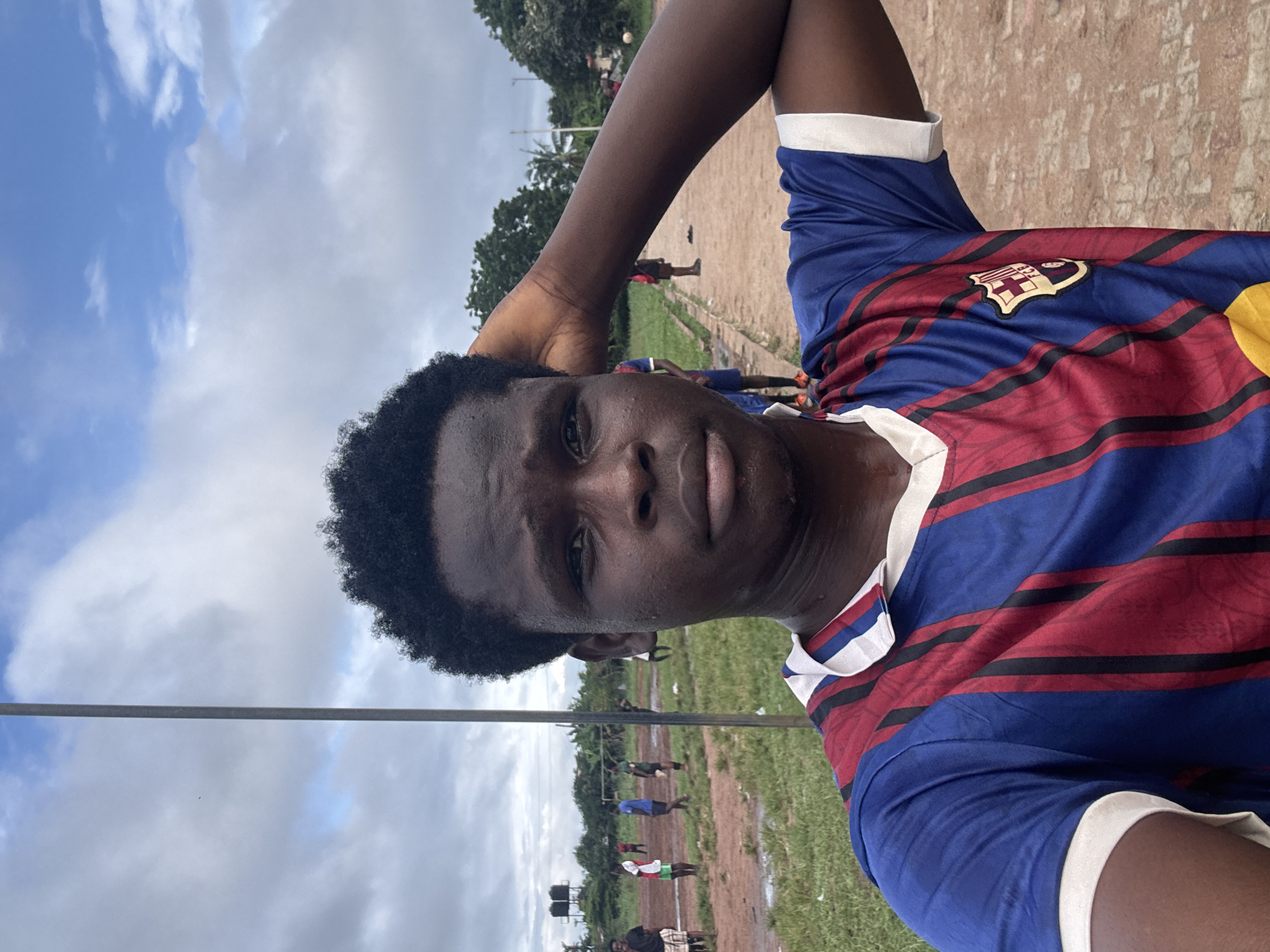 A selfie of a young Black male in the foreground, wearing a red and blue striped soccer jersey with a crest. He has dark, curly hair and is resting his right arm on his head. The background depicts a dirt soccer field with puddles, green grass, and several other individuals, some appearing to be playing soccer. Trees and a cloudy sky are visible. The location is identified as Ihiagwa, Nigeria.