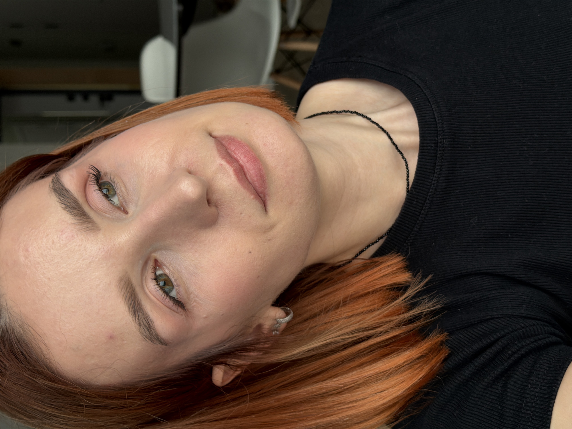 A close-up portrait shows a woman with fair skin and reddish-orange hair. She is wearing a black, ribbed top and a delicate black beaded necklace. An earring is visible in her left ear. Her eyes are light green. She is gazing towards the camera. The background appears to be an interior setting with indistinct furniture and shelving. No specific evidence indicates the location being Chaikovsky, Russia.
