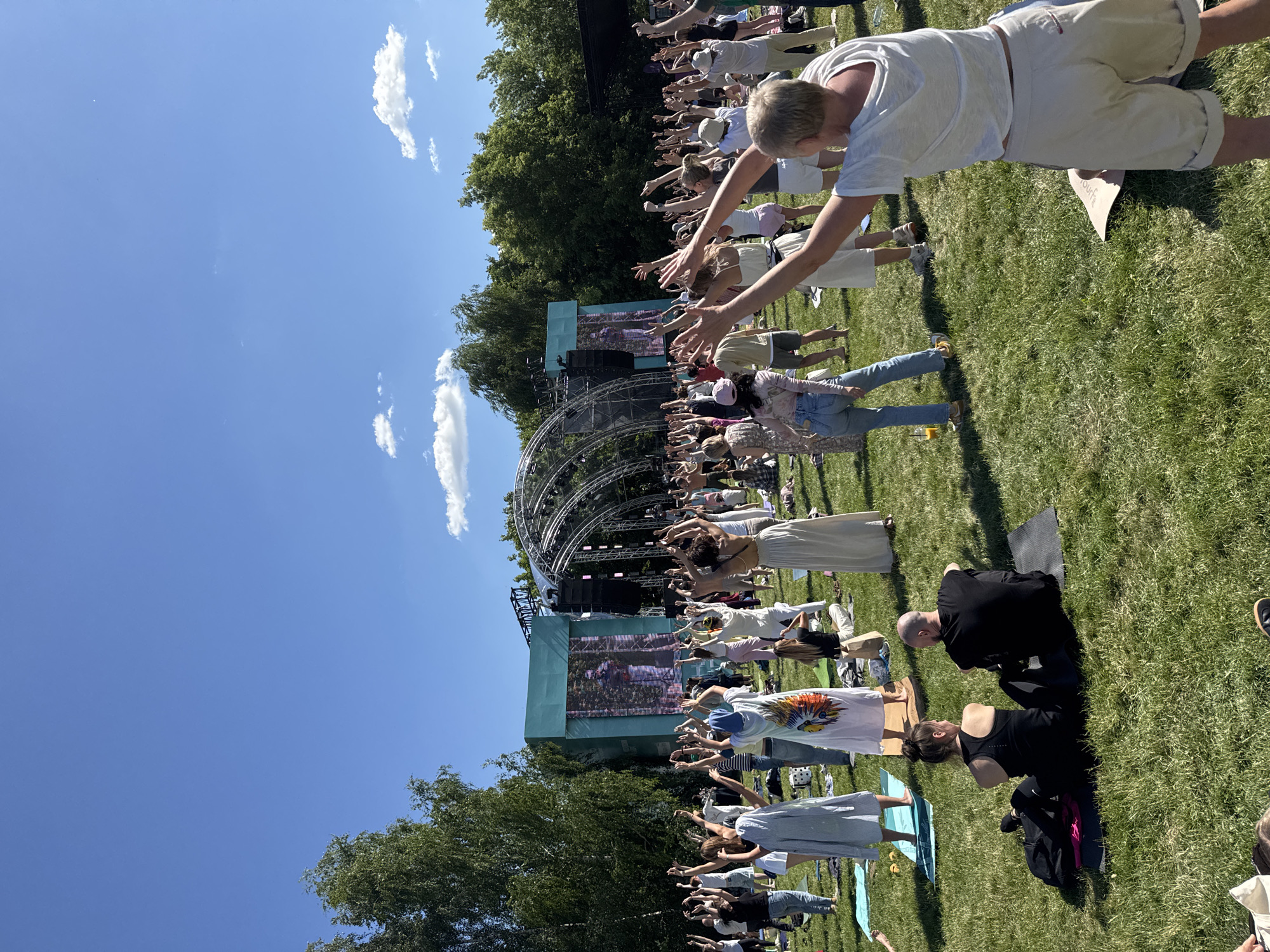 The image captures an outdoor yoga or meditation session taking place on a grassy field under a clear blue sky with scattered clouds. In the background, a stage with a large screen displaying an image of a person is set up, surrounded by metal structures and speakers, suggesting a festival or event setting. A large crowd of people, dressed in casual attire, are participating in the activity, with many of them standing and raising their arms. Some individuals are seated on yoga mats. The environment appears to be a park or open area with trees visible in the distance. The scene suggests a public gathering focused on wellness or entertainment. The presence of a stage and event infrastructure indicates it could be a planned event within a city park.