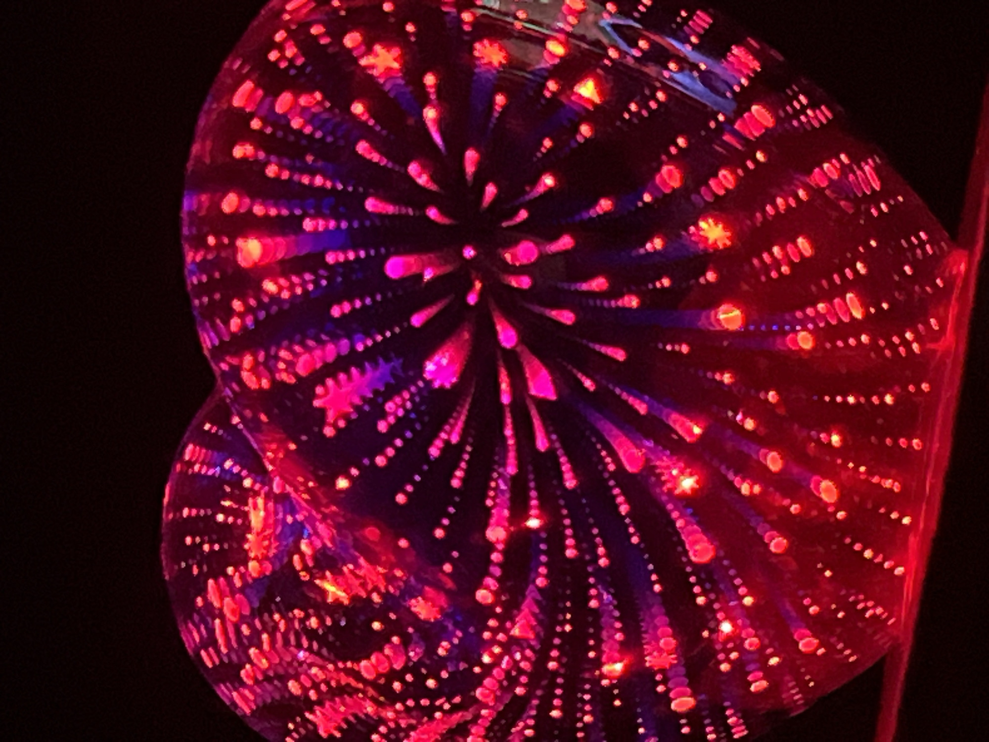 The image shows a close-up, dark shot of a heart-shaped object that appears to be made of glass or crystal. The object is illuminated from within with red and purple lights that create a firework-like pattern of radiating lines and dots. The lights are concentrated in the center of the heart and spread outwards. The background is entirely black, making the illuminated heart the sole focus of the image. The object is placed on a dark surface, with a hint of red light reflecting from below. The image is slightly blurry, giving it an abstract and artistic quality. The specific location in the city of Unknown, Unknown cannot be determined from the visual information provided.