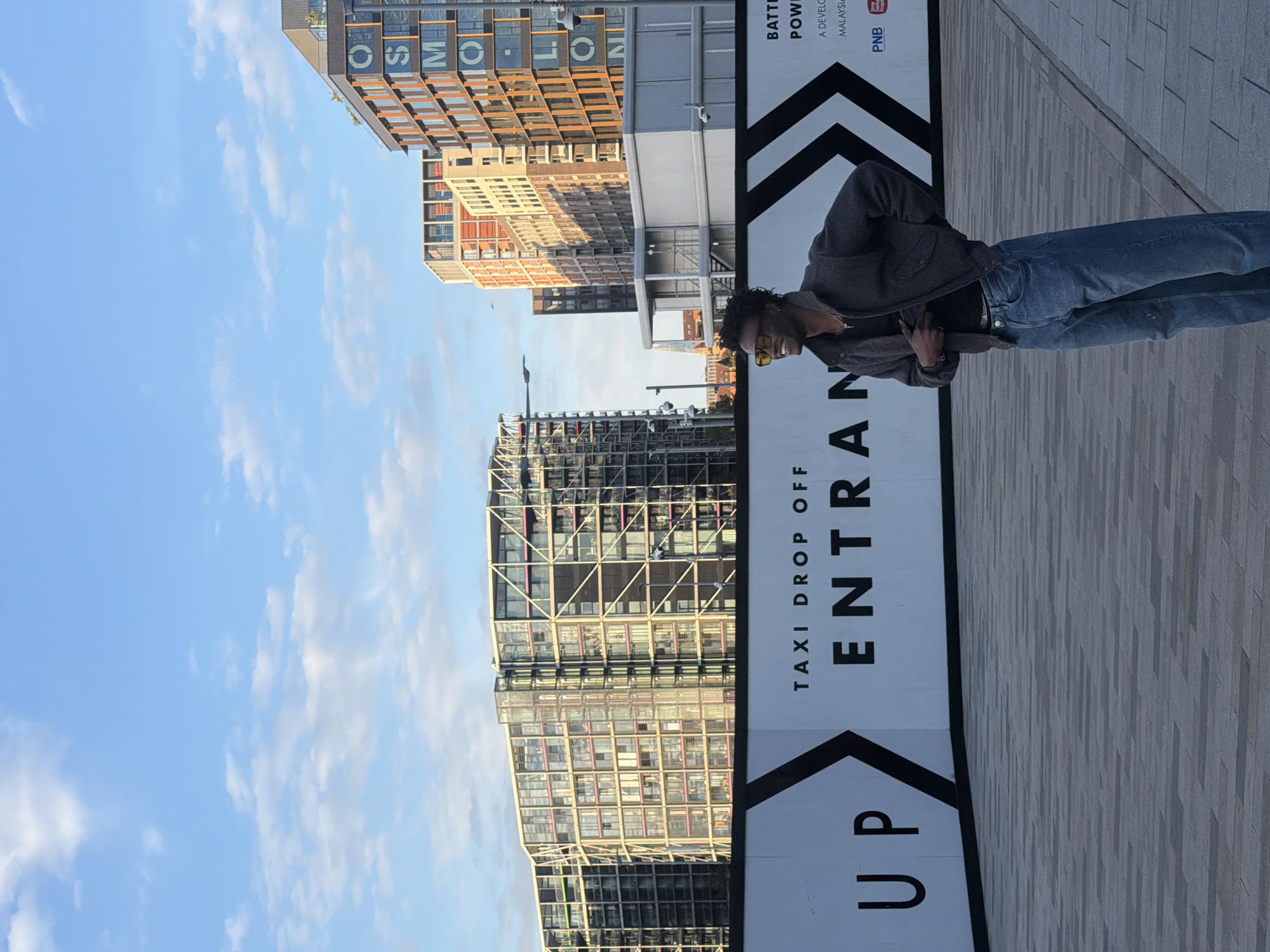 The image shows a person wearing a gray jacket and blue jeans standing on a light gray tiled surface in front of a sign that reads "UP Taxi Drop Off ENTRANCE." The sign is black and white with an arrow pointing upwards. In the background, there are several modern buildings constructed of glass and steel, with one appearing to have large letters spelling out the word "COSMOPOLITAN." The sky is blue with scattered white clouds. Based on the architecture and signage, it is likely that the location is within the city of London, United Kingdom.