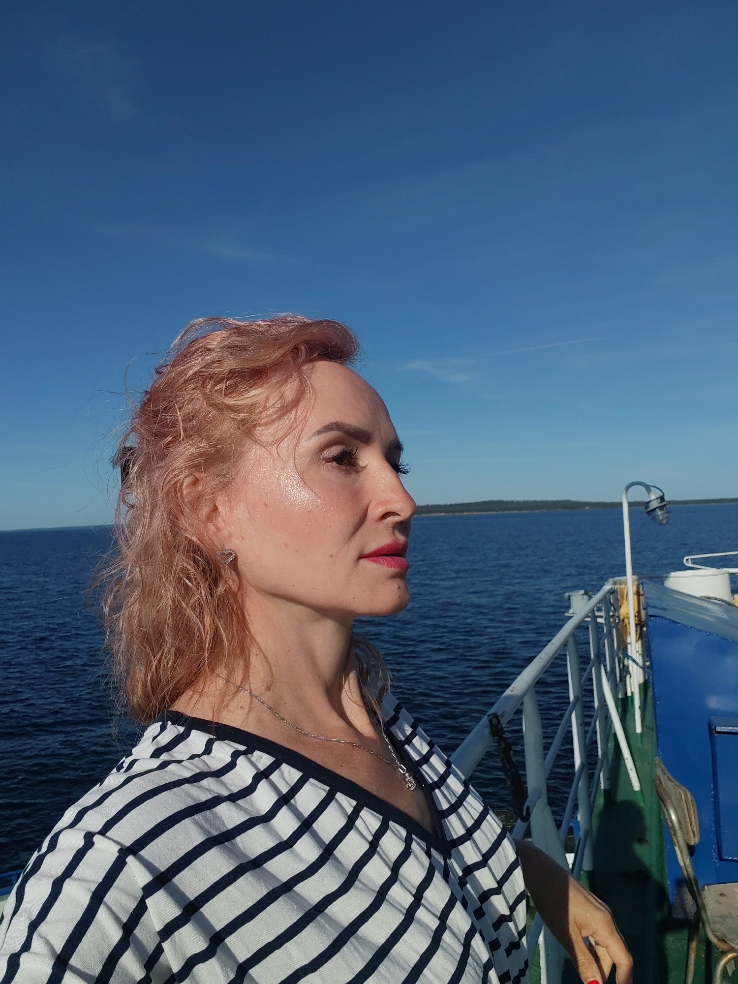 A woman with blonde, wavy hair and fair skin is shown in profile, looking to the right. She is wearing a black and white striped top and a silver necklace. Her makeup includes pink lipstick and mascara. She is standing on the deck of a boat, with the blue water and a hazy shoreline in the background. The sky is clear and blue. A railing and part of the boat's structure are visible in the foreground. The image appears to have been taken on a sunny day. The location is unknown, but the context suggests it is a waterfront or sea-based area.
