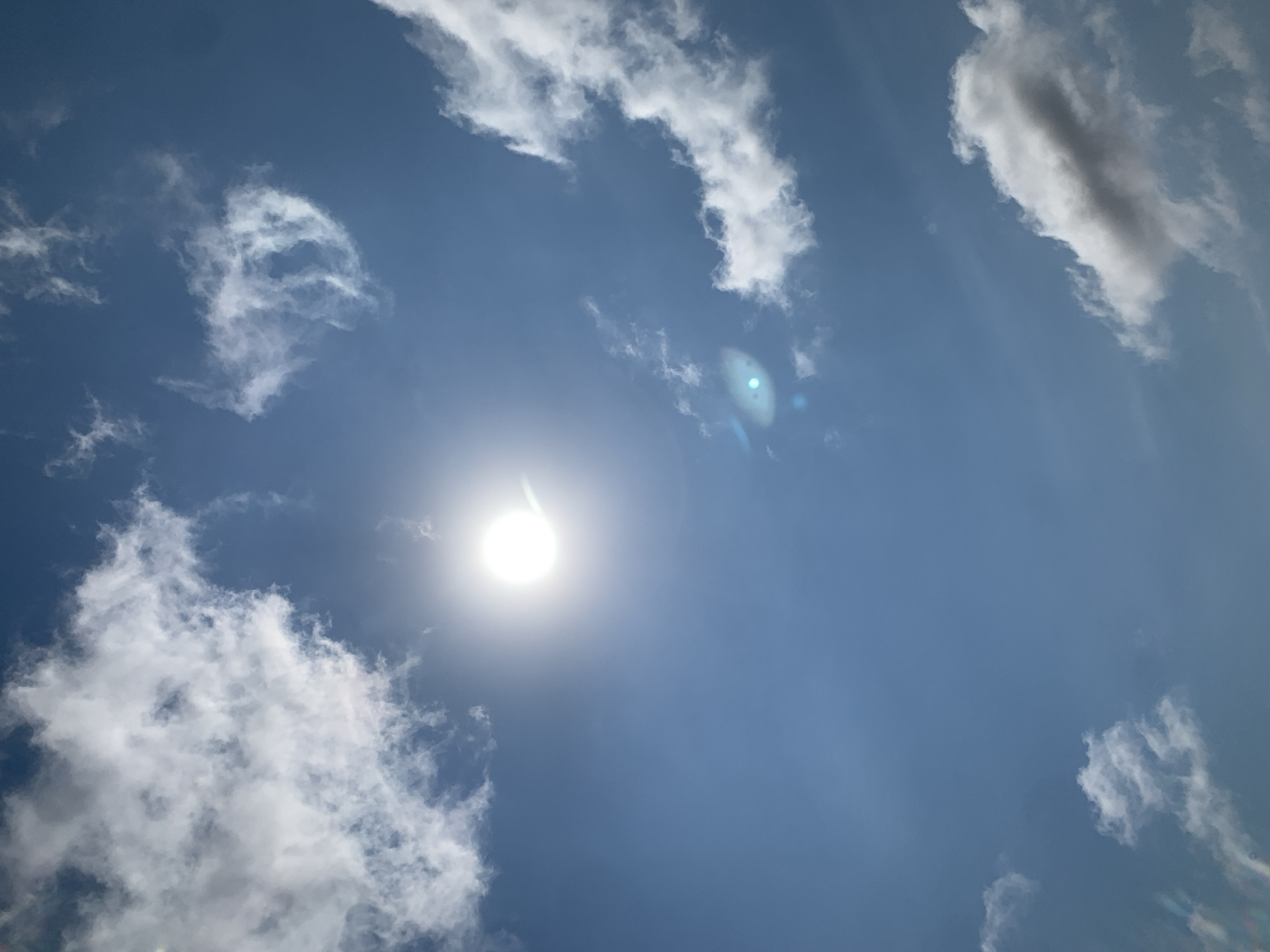 The image is a low-angle shot looking up at a bright, midday sky with scattered white clouds and the sun as the dominant feature. The sun is a brilliant white orb with a visible lens flare extending to the right. Some of the clouds are fluffy and billowy, while others are wispy and thin. The sky is a clear, deep blue. There are no people, buildings, or any other objects visible in the frame. The scene appears to be an outdoor environment, likely a clear day. The location is specified as Khazar Raion, Azerbaijan.