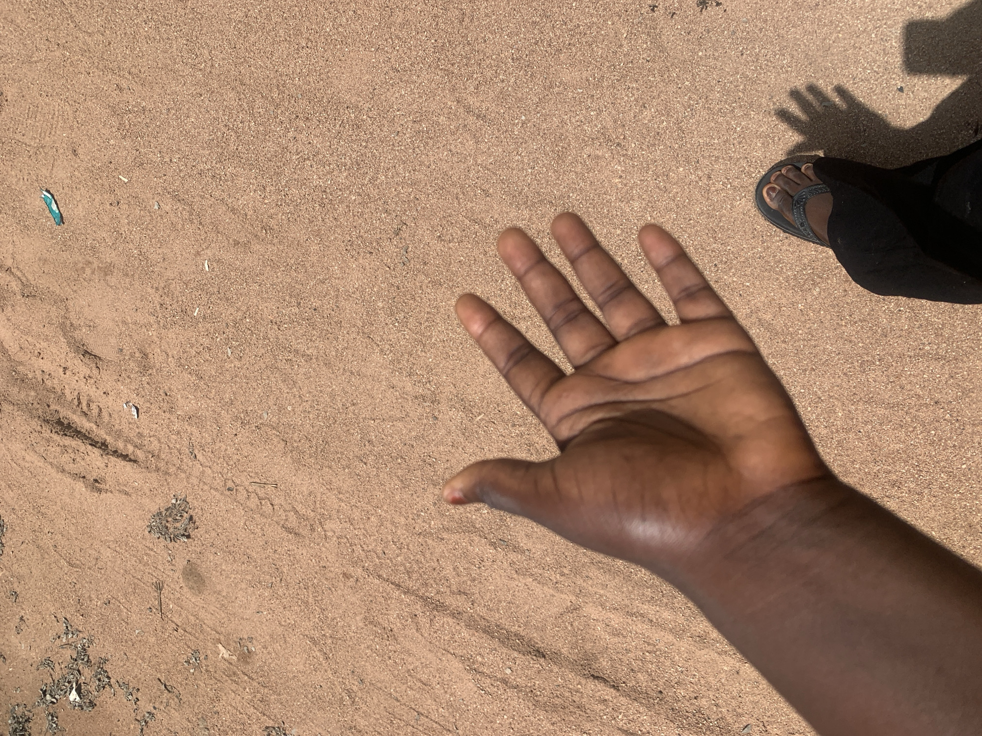 A close-up, overhead shot captures an outstretched hand and a person's foot in dark pants and a flip-flop, casting a shadow on dry, sandy ground in Jauro Yinu, Nigeria. The sand is light brown and speckled with small pebbles and debris, with some faint tire tracks visible. A small, bright blue and white object, possibly a piece of trash, lies in the upper right quadrant of the frame. The hand, with dark skin, is open with fingers slightly spread, and a tiny red mark is visible on the tip of the thumb. The scene suggests an outdoor environment, possibly a dirt road or a cleared area in a rural or semi-urban setting within Jauro Yinu.