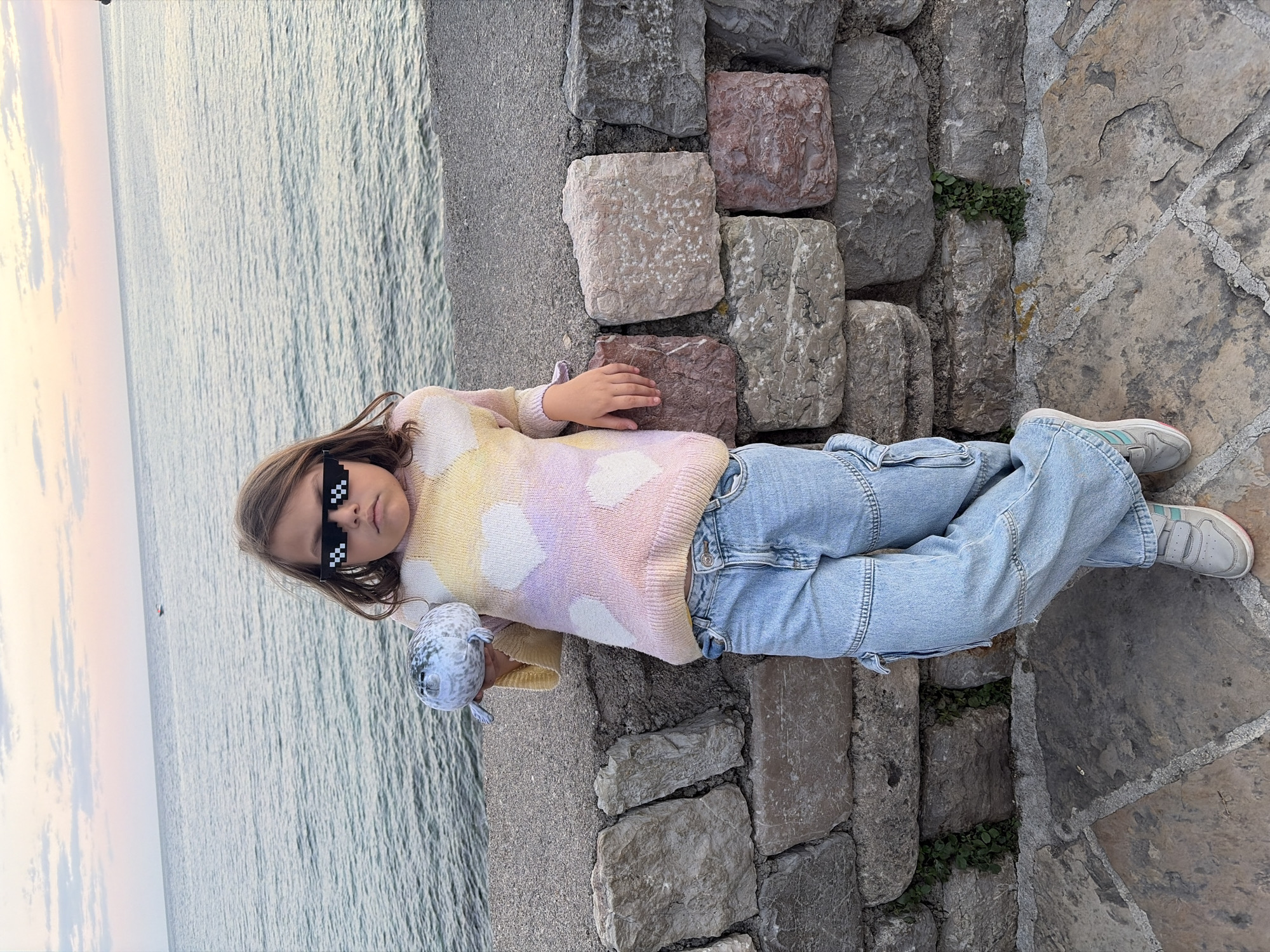A young girl wearing pixelated sunglasses, a pastel-colored sweater, and light blue jeans lies on a stone structure next to the sea. The girl is holding a toy seal. The stone structure is made of various sized stones. The background features the sea and a sky with soft sunset colors. The scene is likely in Budva, Montenegro, given the coastal environment and stone construction.