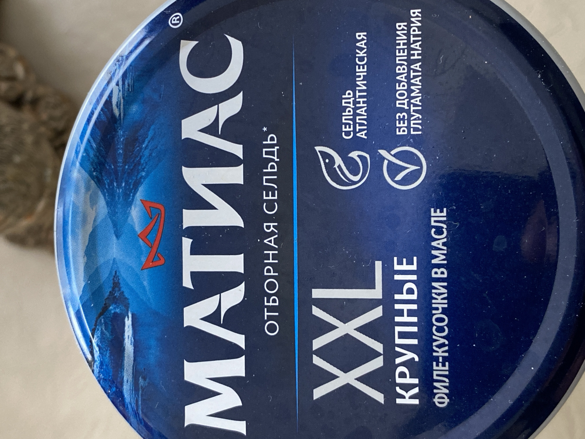 The image shows the top of a round, blue can with white text in Russian. The text reads "МАТИАС" (MATIAS) in large, vertically oriented letters on the left side, followed by "ОТБОРНАЯ СЕЛЬДЬ*" (Selected herring*). Below this is "XXL" and "КРУПНЫЕ" (Large), and then "ФИЛЕ-КУСОЧКИ В МАСЛЕ" (Fillet pieces in oil).
To the upper right of the main text, there are descriptions of "СЕЛЬДЬ АТЛАНТИЧЕСКАЯ" (Atlantic herring) and "БЕЗ ДОБАВЛЕНИЯ ГЛУТАМАТА НАТРИЯ" (No added sodium glutamate), accompanied by icons. At the top left, there is a registered trademark symbol. The can has a dark blue lid. A landscape featuring a castle is printed on the left portion of the can's side.