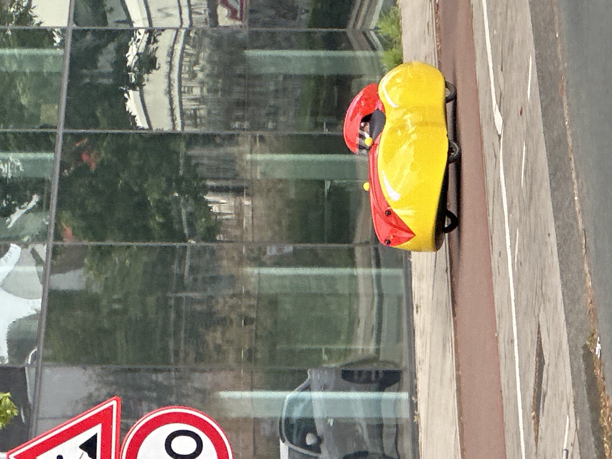 The image shows a yellow and red toy car situated on the edge of a walkway or sidewalk, partially bordering a building with reflective glass windows. The reflection in the windows reveals trees, buildings, and other elements of an urban environment. In the lower portion of the image, traffic signs are visible, further suggesting an urban setting. Given the presence of Dutch traffic signs and other visual clues, the location is likely Amsterdam, Netherlands. The image does not depict any people or direct actions. The key objects are the toy car, the building facade, and the traffic signs. The environment is a cityscape with a mix of modern architecture and natural elements.