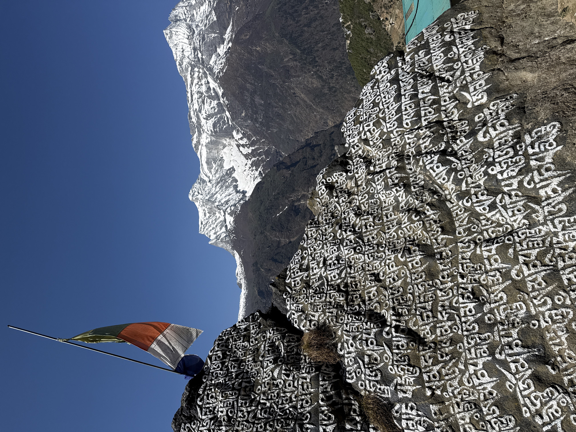 A mountain in Namche, Nepal, is shown to the left, and a large rock with Om Mani Padme Hum writings is on the right. A flagpole stands in front of the rock with a prayer flag. The sky is clear and blue.