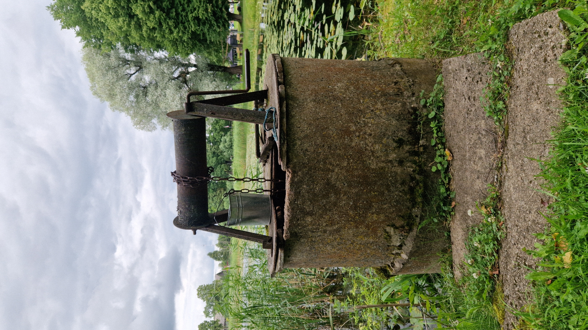 This is a full shot of an old, weathered concrete well structure situated outdoors. The well has a rusty metal mechanism for drawing water, with a bucket hanging from a chain. The well is surrounded by lush green grass, reeds, and a body of water with lily pads. In the background, trees and a cloudy sky are visible. The scene appears to be in a rural or park-like setting. While the specific location in Priekule, Latvia is mentioned, there are no visible landmarks or indicators within the image to confirm this. There are no people or animals present in the image.