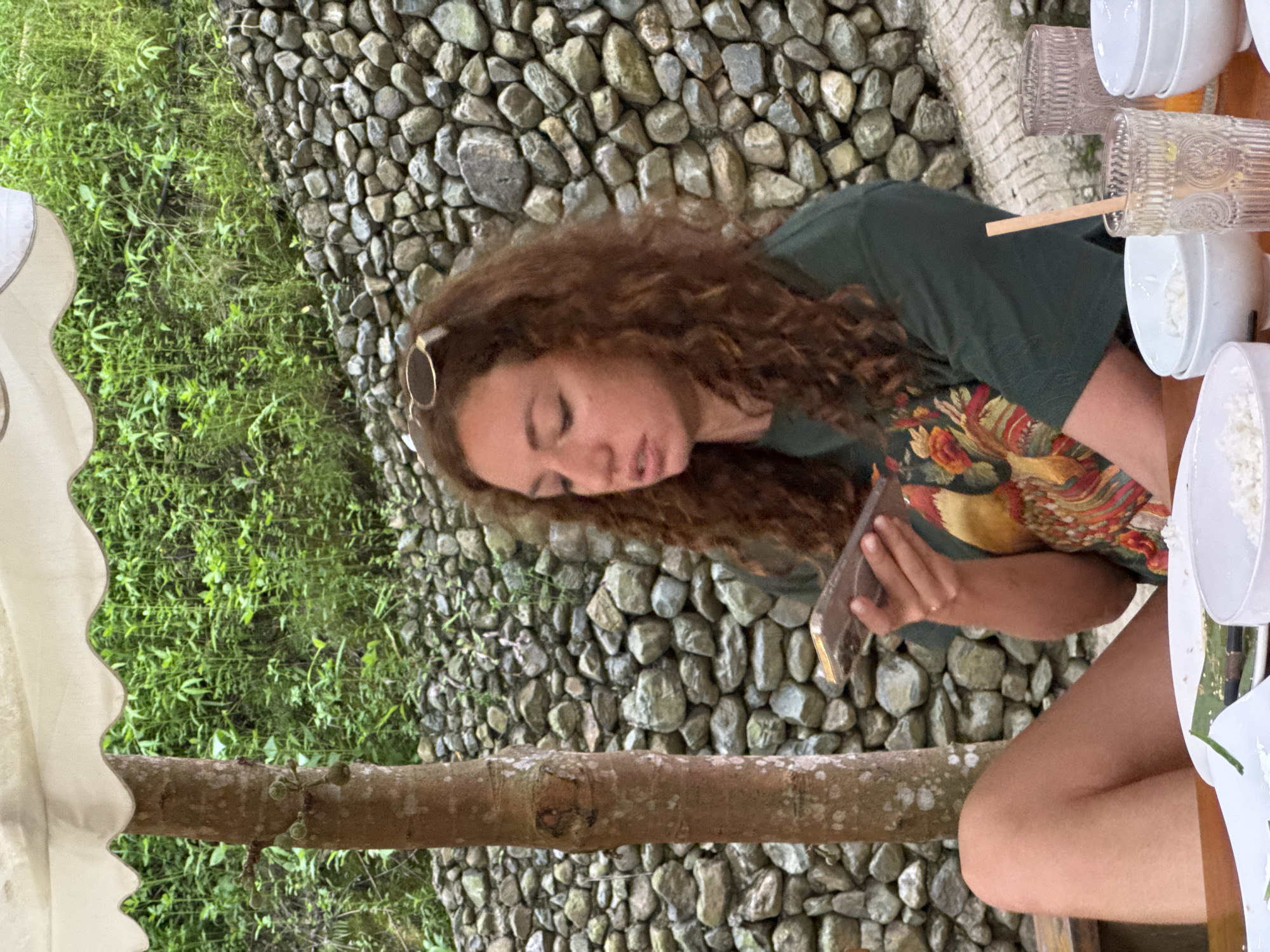 A woman with long, curly brown hair sits at a table outdoors, looking down at a mobile phone she holds in her left hand. She wears a dark green t-shirt with a colorful, patterned design on the front, featuring what appears to be a bird or rooster. Her legs are crossed, and she is positioned to the left of the frame. In front of her are white bowls filled with rice, and to the right, clear glasses, one with a straw and what looks like a drink. The background consists of a rustic stone wall, lush green vegetation, and a tree trunk to the left of the woman. The setting appears to be a restaurant or outdoor dining area in Cổ Lũng Commune, Vietnam, suggested by the natural, tropical environment.