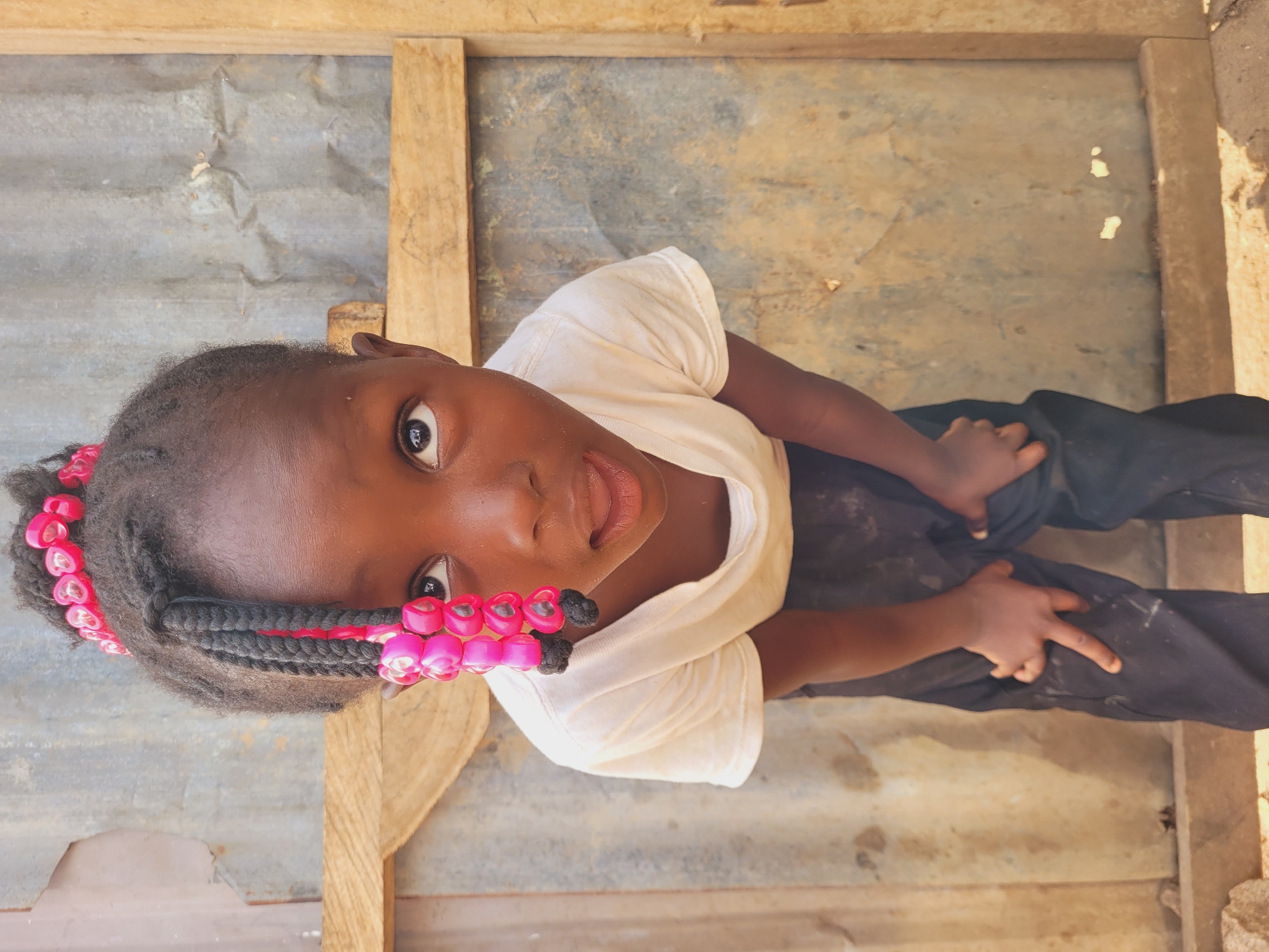 An overhead, medium shot depicts a young African child, likely female, with dark skin and hair braided with pink beads. She is wearing a light-colored t-shirt and dark-colored trousers. The child is looking up directly at the camera. The background is a corrugated metal wall and a wooden frame. The location is assumed to be Bukuru, Nigeria, based on the prompt. There is no information available to confirm that.