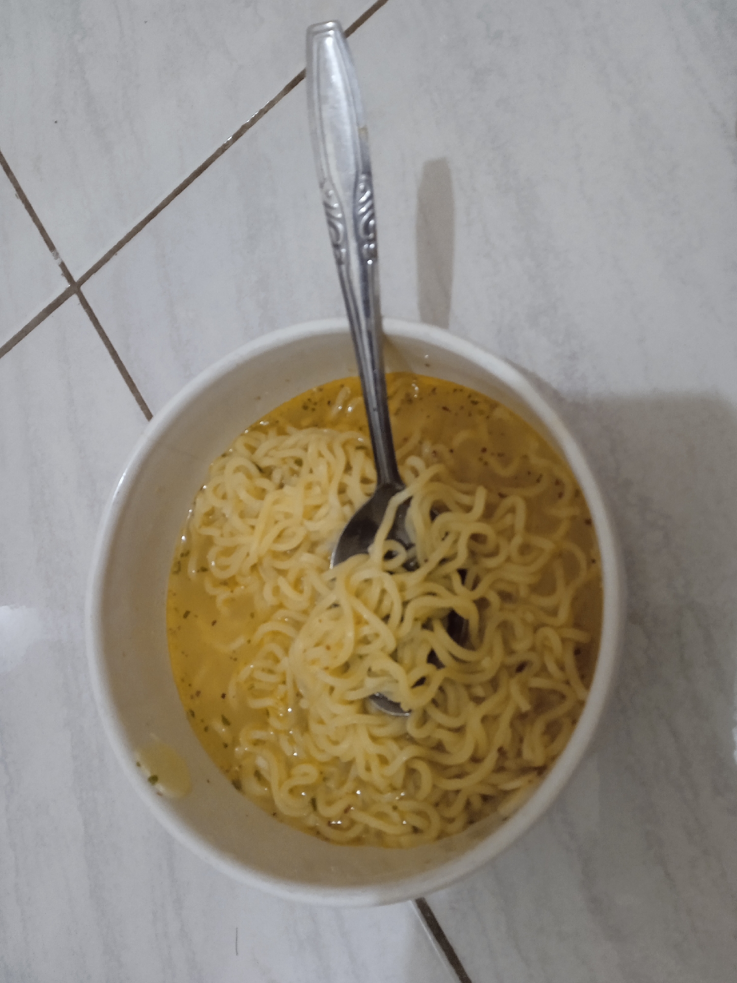 A white, round bowl containing a serving of instant noodles in a yellow, opaque broth is positioned centrally on a light-colored tiled floor in Brangsong, Indonesia. A silver-toned metal spoon, featuring an embossed decorative pattern on its handle, is inserted into the noodles and broth, with a portion of the handle extending upwards from the bowl. The noodles are numerous and light yellow, exhibiting a characteristic curly texture. Small dark specks, consistent with dried herbs or seasoning, are visible within the broth. The tiled floor comprises light-colored, possibly off-white or light grey, square or rectangular tiles with darker, narrow grout lines forming a grid pattern. A soft shadow is cast by the bowl and spoon towards the right side of the image, indicating a primary light source originating from the left. No individuals are visible in the frame.