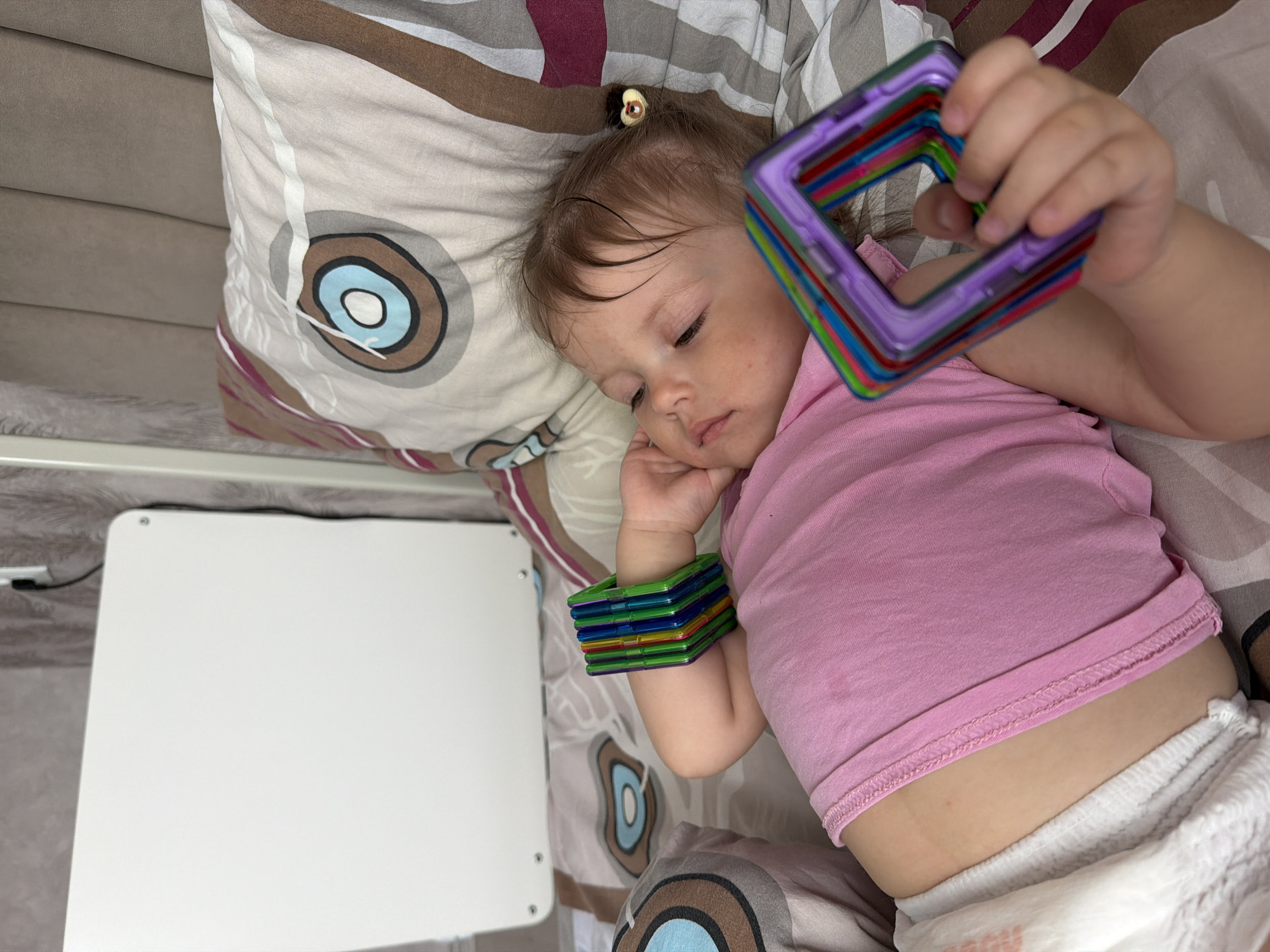 A baby girl, clad in a pink t-shirt and white diaper, is lying on a patterned pillow. She has magnetic toys on her wrist and in her hand. Next to her is a white square table. The location is unspecified and there is no evidence to indicate that this image was taken in Reshetnikovo, Russia.
