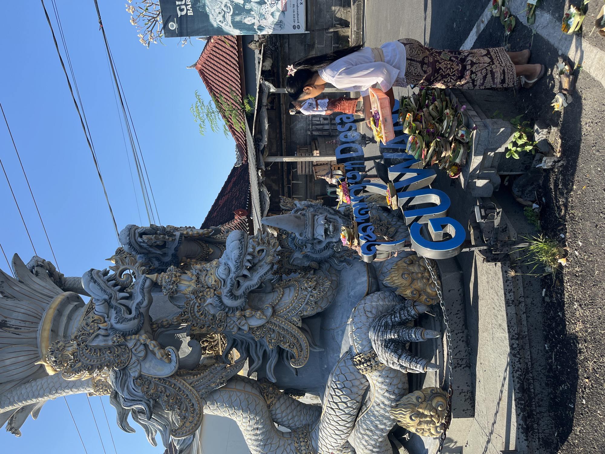 The image depicts a street scene in Sukawati, Indonesia, featuring a prominent dragon statue. A young woman, dressed in traditional Balinese attire, is arranging offerings at the base of the statue. The offerings, consisting of woven leaf baskets filled with flowers and other items, are meticulously placed on a stone ledge and the ground. In the background, several structures with red tiled roofs are visible, along with street signs and utility wires. The overall atmosphere suggests a cultural or religious setting.