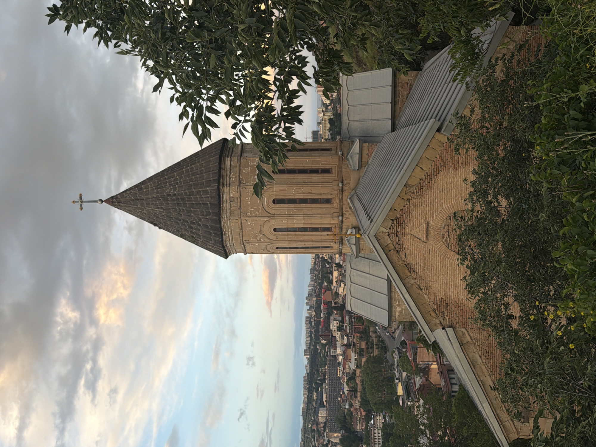 The image depicts a tall, brick structure with a conical roof and a cross at its apex, situated in Tbilisi, Georgia. The building appears to be a church or religious building, surrounded by trees. The cityscape of Tbilisi stretches out in the background. The sky is overcast with clouds.