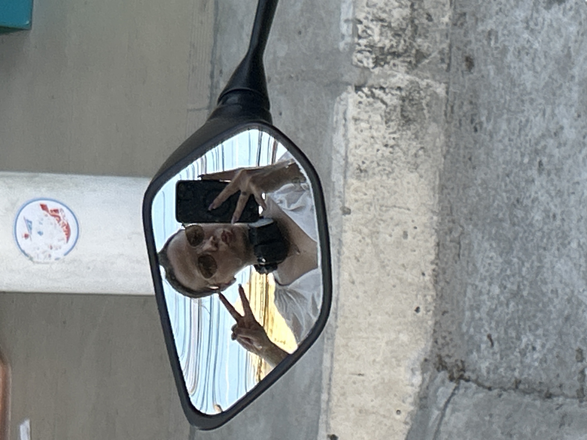 A woman is reflected in a motorcycle's side mirror, taking a selfie. She is wearing sunglasses and a white top, with headphones around her neck. She is making a peace sign with her left hand and holding a smartphone with her right. The reflection shows a background of horizontal blue and white stripes, suggesting a striped awning or fabric. The surrounding environment appears to be outdoors, with a concrete wall visible in the foreground and background. The image is likely taken in Baan Hua Thanon, Thailand, based on the context provided.