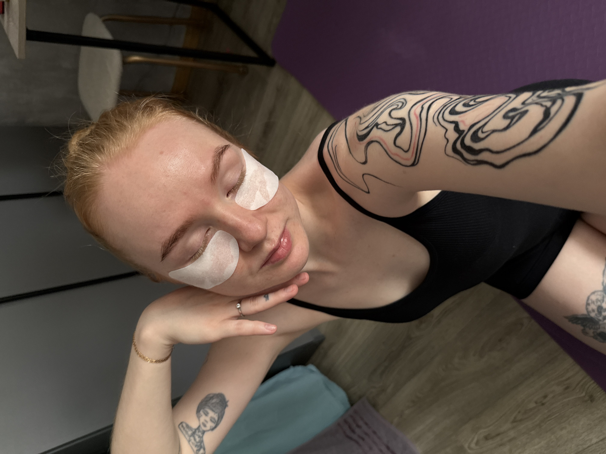 A young, fair-skinned woman with red hair tied up in a bun is looking down with her eyes closed, wearing white under-eye patches. She has tattoos on both arms, one on her left arm depicting a woman's face, and a swirling abstract design on her right arm with black and red lines. She is wearing a black tank top and appears to be sitting on a purple yoga mat on a wooden floor. The background is blurred and shows a closet and a stool with a light-colored seat and dark legs. The image seems to have been taken indoors, possibly in a residential setting, and there is no indication of the city of Chaikovsky, Russia, within the visual information provided.