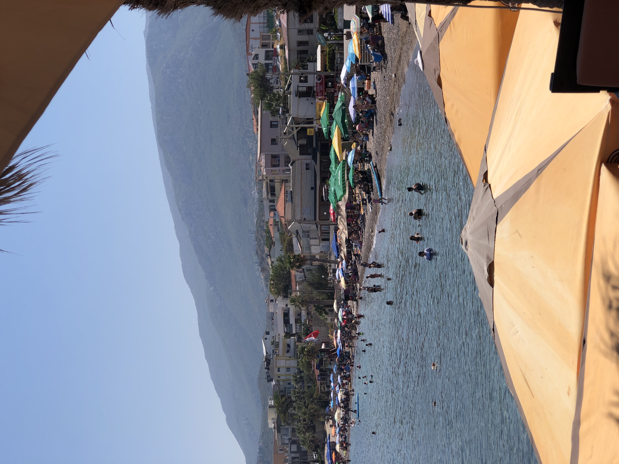 The image captures a bustling beach scene in Karaburun, Turkey, on a clear, sunny day. The foreground is dominated by the tops of several large, orange and brown umbrellas, suggesting a cafe or seating area overlooking the water. Beyond the umbrellas, the sea is filled with people swimming and enjoying the water. The shoreline is lined with a crowded beach, populated by numerous individuals relaxing under a variety of colorful umbrellas, including green, yellow, blue, and striped ones. Buildings, likely hotels or shops, are visible behind the beach, extending up the gentle slope of the land. In the background, a large, verdant mountain range rises majestically under a pale blue sky. A Turkish flag is prominently displayed on a pole near the buildings. The overall impression is one of a vibrant and popular summer destination.