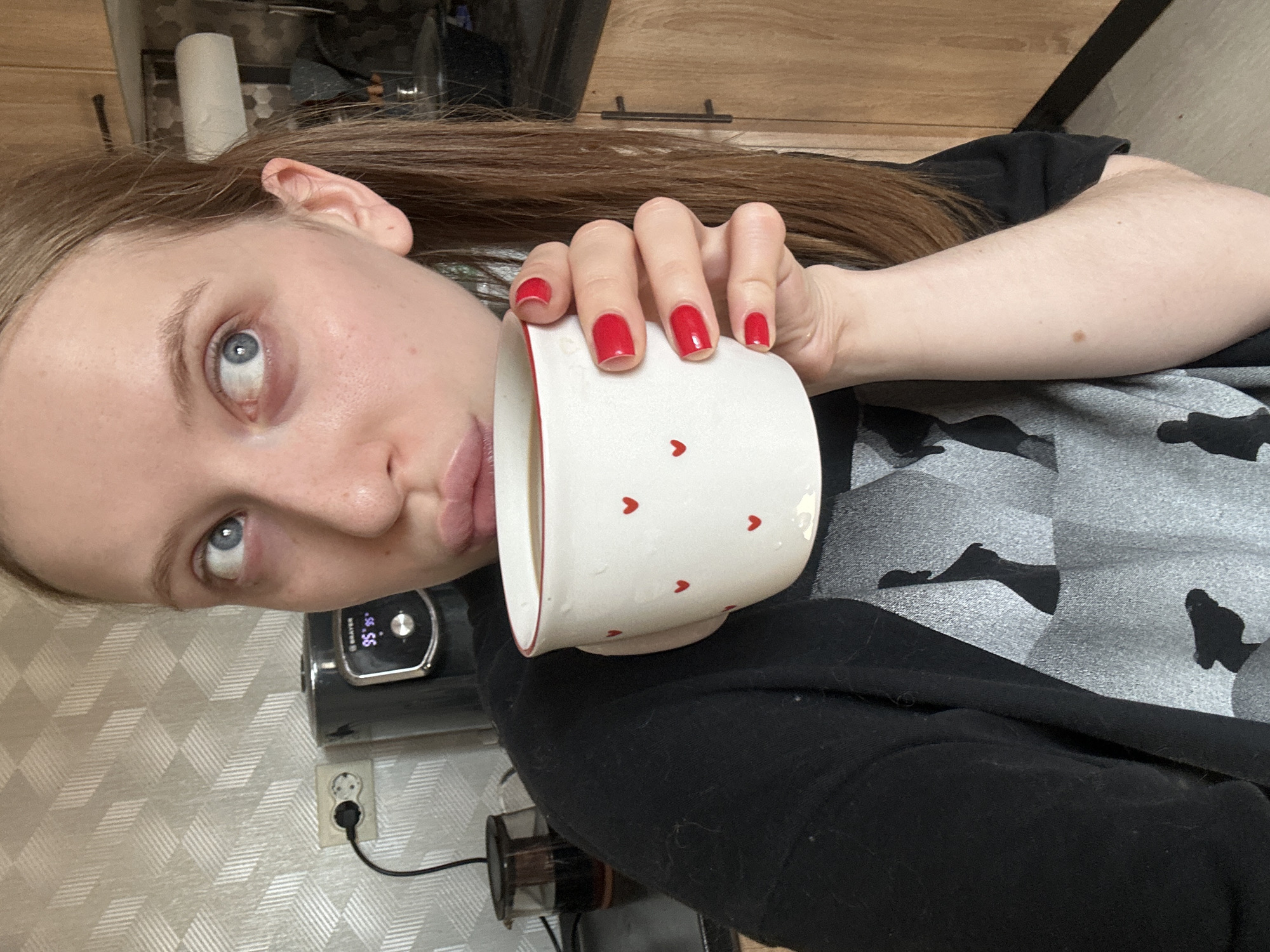 A fair-skinned woman with blue eyes and light brown hair is holding a white mug with red heart designs, seemingly drinking from it. She has red nail polish on her fingernails. Her clothing consists of a gray patterned t-shirt covered by a black garment. The environment appears to be an indoor kitchen setting, including a coffee maker and a patterned wall with an electrical outlet. The location is not discernible from the image alone.