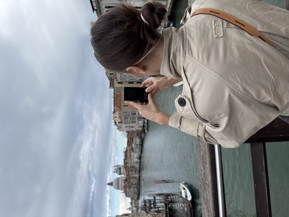 An overhead view captures a person from behind, positioned on a bridge or elevated structure, photographing the Grand Canal in Venice, Italy, on an overcast day. The subject, appearing female, wears a beige trench coat and has brown hair tied in a bun with a pink hair tie. A light brown leather strap crosses her right shoulder. She holds a black smartphone or compact digital camera vertically with both hands, positioned as if taking a photograph. In the midground, the Grand Canal flows with greenish water, lined by multi-story historic Venetian buildings featuring traditional orange-tiled roofs and light-colored facades. A white motorboat with blue trim is docked on the left side of the canal, near wooden mooring poles. In the background, the distinctive large dome and smaller domes of the Basilica di Santa Maria della Salute are prominently visible on the left side of the canal. The sky is entirely covered with gray and white clouds.