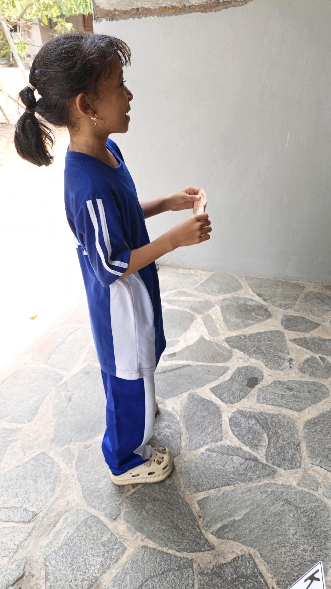 A young girl with dark hair styled into two pigtails, wearing a blue and white t-shirt and blue pants, stands on a stone pathway. She has a golden hoop earring in her left ear. In her hands, she holds a small, light-colored object, possibly a piece of paper or candy wrapper. The background consists of a plain gray wall and some greenery visible to the left. The pathway is made of irregularly shaped gray stones. This scene appears to be outdoors, likely in a residential or public area within Jepara, Indonesia.