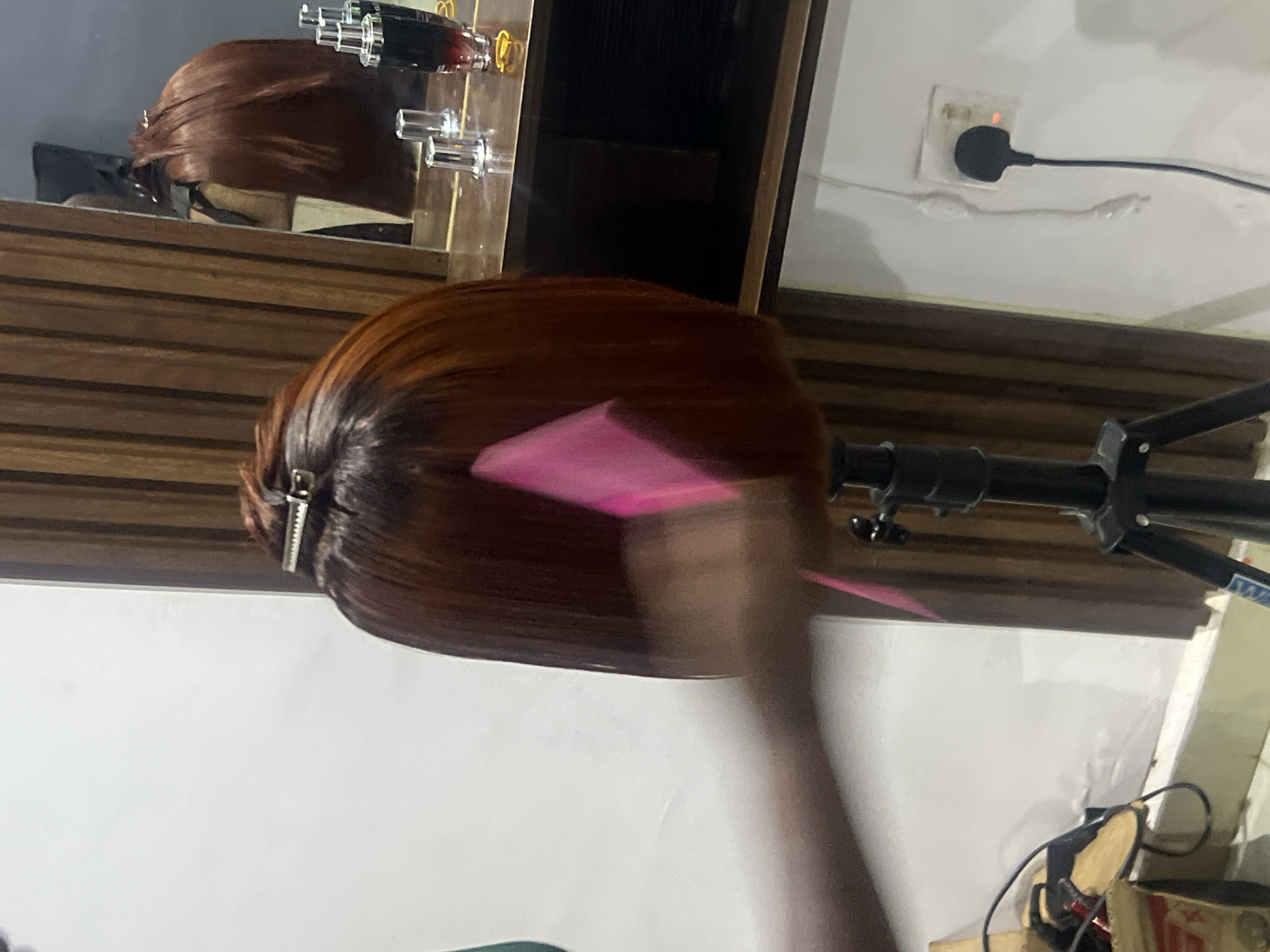 The image depicts a close-up of a mannequin head with reddish-brown hair, styled in a bob. The hair is secured with a silver clip at the crown. A pink comb is in motion, blurred due to being moved, positioned near the side of the mannequin's head. The mannequin head is mounted on a black tripod stand. In the background, a wooden-paneled wall is visible, and to the right, a mirror reflects more of the mannequin's hair, as well as a countertop with some bottles and accessories. The overall setting appears to be a hair salon or a similar professional grooming space. There is no discernible connection to Gura Top, Nigeria, in the provided visual information.