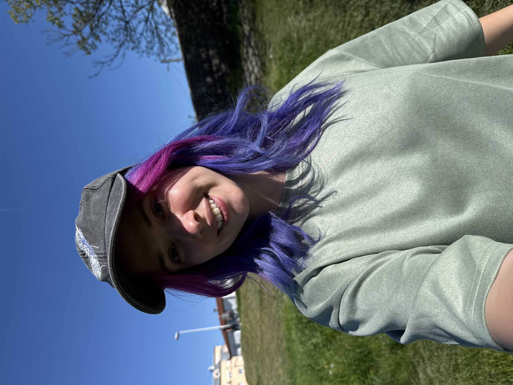 A selfie-style photo, rotated ninety degrees clockwise, captures a young woman outdoors on a bright, sunny day. The woman is the primary subject, shown from the chest up, smiling cheerfully into the camera. She has striking, vibrant hair, predominantly purple with noticeable streaks of pink. She is wearing a dark grey baseball cap with a light-colored, possibly white or light blue, embroidered design on its front brim. Her attire includes a light sage green, long-sleeved top with a slightly textured fabric.
Her smile is broad, revealing her teeth, and her expression conveys happiness or enjoyment. Direct sunlight illuminates her face, creating a mix of bright highlights and soft shadows.
The background features a clear, vivid blue sky taking up a significant portion of the frame, indicating excellent weather. Below the skyline, glimpses of lush green grass and the dark trunks and green foliage of trees are visible. In the distance, on the right side, there are hints of light-colored buildings and a slender street light pole, suggesting an urban or suburban park setting. No visible text is present in the image. The scene takes place in Zagreb, Croatia.