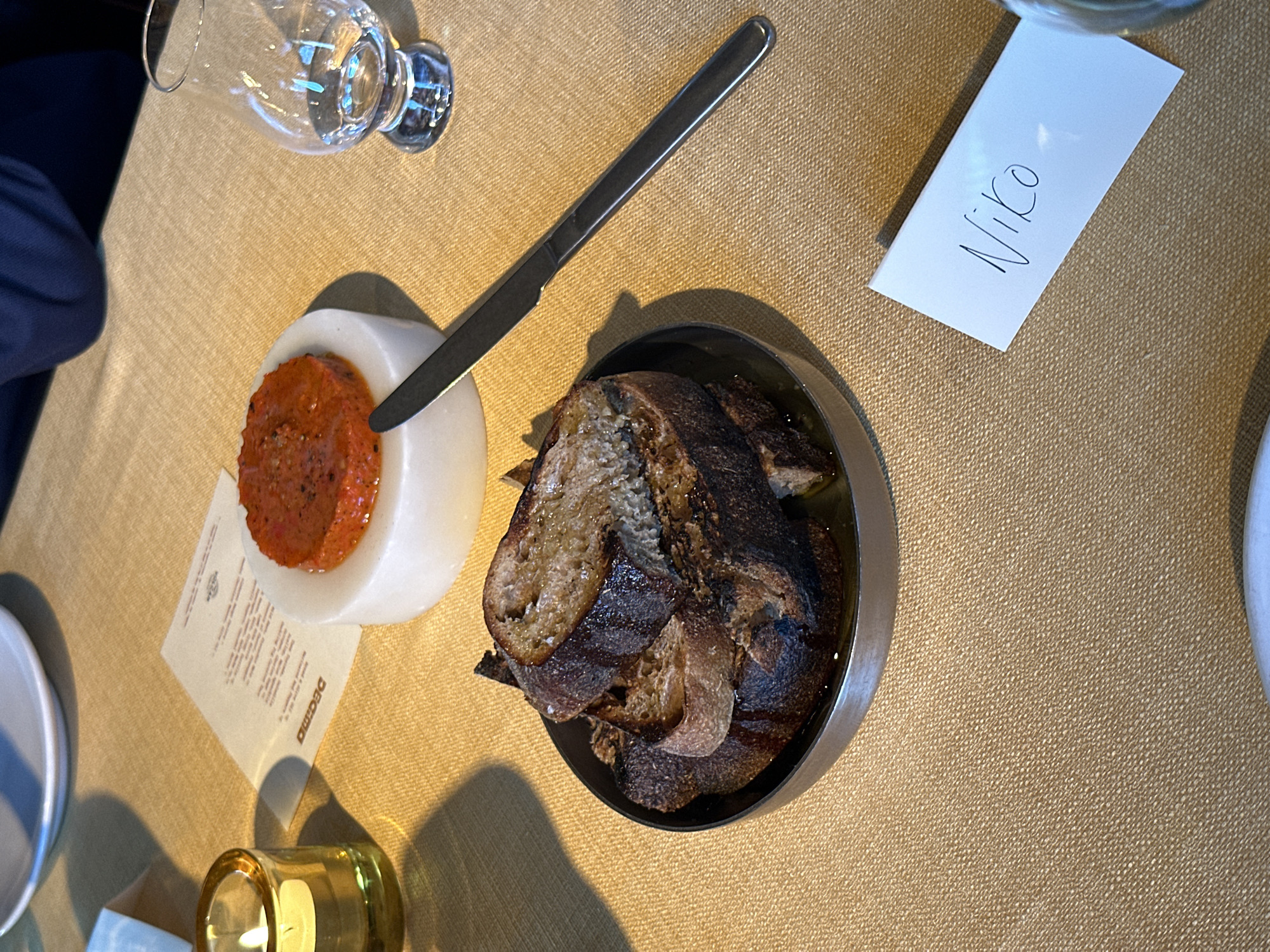 The image depicts a table setting, likely in a restaurant. A bread plate with dark, crusty bread slices sits to the right. A white pedestal holds a red spread with a knife resting on it. A glass and a white name card with "Niko" written on it are also visible. The surface is a textured tan color, possibly a tablecloth. A menu or printed card is placed near a small, candle holder. The scene suggests a formal or semi-formal dining experience. The city of London, United Kingdom, is not discernable from the provided image.
