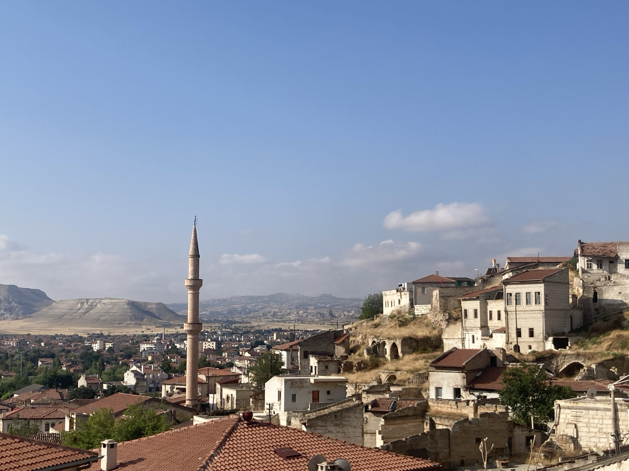 The image depicts a panoramic view of Avanos İlçe Merkezi, Turkey, on a clear day with a bright blue sky. In the foreground, terracotta tiled rooftops of various buildings are visible. A prominent minaret, characteristic of Islamic architecture, stands tall to the left of the frame, its slender structure reaching towards the sky. The middle ground showcases a cluster of buildings with light-colored facades, some appearing to be carved into the hillside. These structures exhibit architectural styles typical of the region, with windows and doorways. Beyond the immediate settlement, a vast expanse of dry, undulating terrain stretches towards the horizon. In the distance, a large, layered rock formation dominates the landscape, its contours weathered by time. The overall scene conveys a sense of historical depth and a tranquil, arid environment. No people are visible in the image.