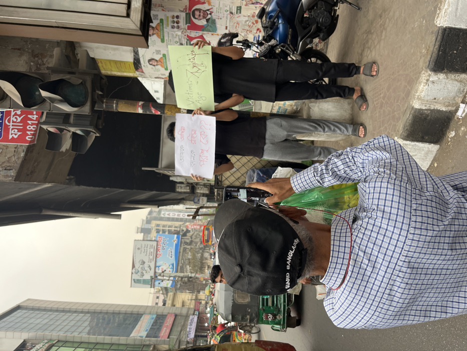The image depicts a street scene in Dhaka, Bangladesh, capturing a moment of public awareness or protest. In the foreground, facing away from the viewer, an older man with grey hair wears a blue and white checkered shirt, a black baseball cap inscribed with "GUARD BANGLA," and red headphones around his neck. He is actively holding a black smartphone vertically with both hands, appearing to record or photograph the scene ahead. A green plastic bag is held in his left hand.
Directly in front of him, on a concrete sidewalk marked by a black and white striped curb, stand two young men whose faces are obscured by protest signs they hold. The man on the left wears a black t-shirt and grey sweatpants with grey sandals, holding a white sign with red Bengali text which translates to "Today Omiya, Mansu, Roima, tomorrow who??". The man on the right wears a black t-shirt and black sweatpants with black sandals, holding a light green sign with black and red English text that reads "Victims: Age - 7 years". A dark blue motorcycle is parked behind the second protester.
To the right, a dark grey traffic light fixture with three lights is mounted vertically on a pole. The street to the left shows ongoing traffic, with a green three-wheeled auto-rickshaw (CNG) partially visible. The background reveals a busy urban environment with multiple multi-story commercial buildings. Numerous large billboards with advertisements, including one for "Auro," are visible. A red sign with Bengali text "ফাহিম" (Fahim) and a phone number (01814-...) is prominently displayed on a wall. The sky above is overcast or hazy.