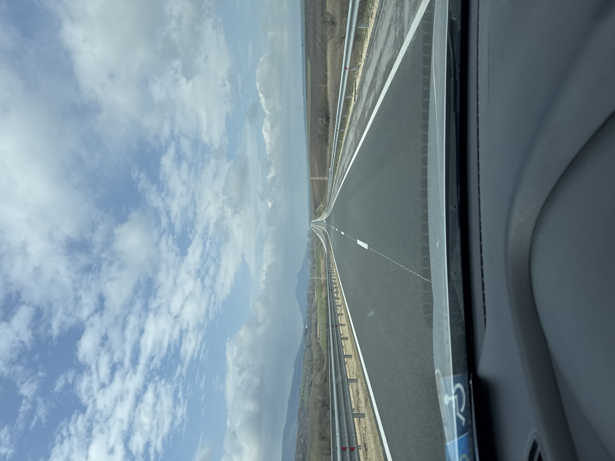 The image shows a view from inside a vehicle traveling on a highway in Gradishte, Bulgaria. The road is asphalt and divided by white lines, with metal guardrails along the sides. The landscape includes fields and distant mountains under a sky with scattered clouds. The view is partially obstructed by the vehicle's interior, including the door and possible window frame. A handicapped symbol is visible on the door.