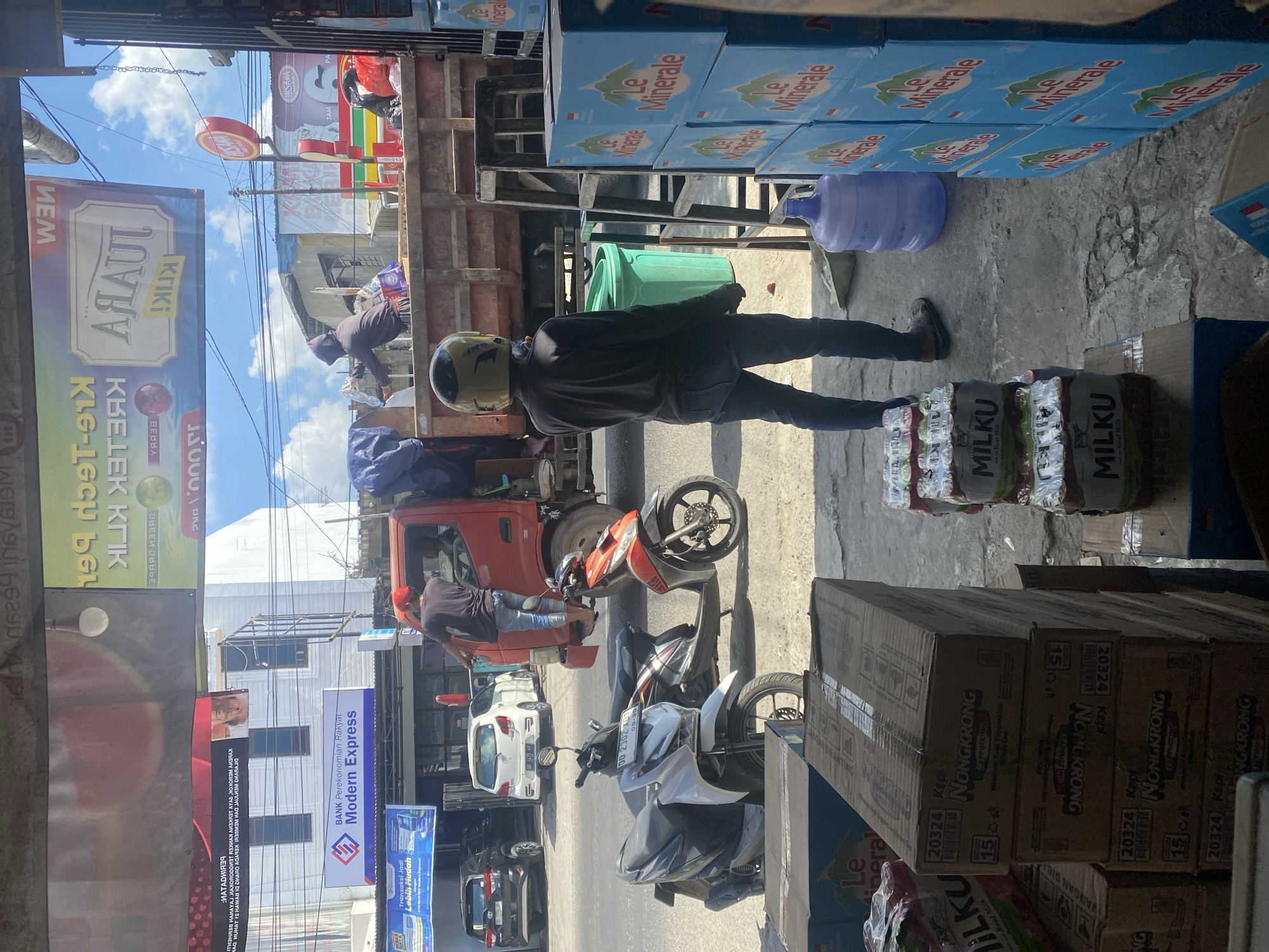 The image depicts a street scene in Bulukumba, Indonesia, likely near a marketplace or shop. In the foreground, stacks of boxed beverages, specifically "Nongkrong Cappuccino" and "Le Minerale" bottled water, are visible. A person wearing a dark long-sleeved shirt, jeans, and a yellow helmet stands facing away from the camera, holding a phone. To the left, two scooters are parked on the street. Behind them, a red truck is parked, with two individuals in the process of loading or unloading items. A person in a red cap is standing on the back of the truck. In the background, buildings, signage, and a bright blue sky with some clouds are present. One sign reads "BANK Perekonomian Rakyat Modern Express". The overall impression is that of a busy commercial area.