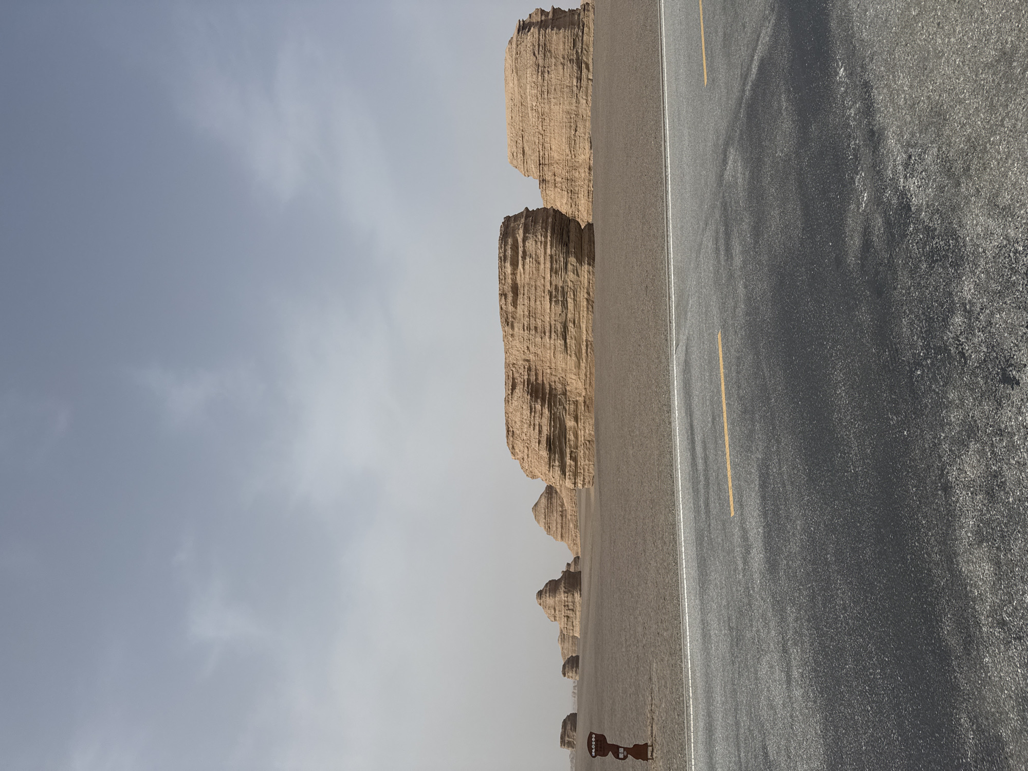 The image, oriented horizontally, depicts a remote desert landscape featuring unique geological formations, an asphalt road, and an expansive sky. The upper portion of the frame is dominated by a pale, overcast sky, ranging from light grey to a muted blue, with subtle cloud patterns indicating diffused daylight.
On the left side, distinctive Yardang landforms rise from the flat desert terrain. These eroded rock structures, appearing in shades of light brown and beige, showcase vertical striations and layers, diminishing in size as they recede towards the distant horizon. This succession of formations suggests a vast, ancient landscape.
A dark grey asphalt road occupies the right side of the image, extending from the foreground into the background. It features a clear white solid line marking the shoulder and a yellow dashed line indicating the center of a two-lane thoroughfare. The road surface exhibits a slight reflectivity. Between the road and the Yardang formations, the ground is a flat, sandy or gravelly desert floor, consistent with the surrounding environment.
In the lower-left corner, a small, dark, stylized sign, possibly shaped like a person, stands near the road edge. It bears some unreadable white text or symbols.
There are no people or vehicles visible, conveying a sense of solitude and vastness. The diffused light suggests a cloudy day, possibly midday or early afternoon. Given the location context of Dunhuang City, China, these formations are characteristic of the Dunhuang Yardang National Park, known for its unique wind-eroded landscapes. The scene evokes a feeling of awe and isolation amidst a grand natural setting.