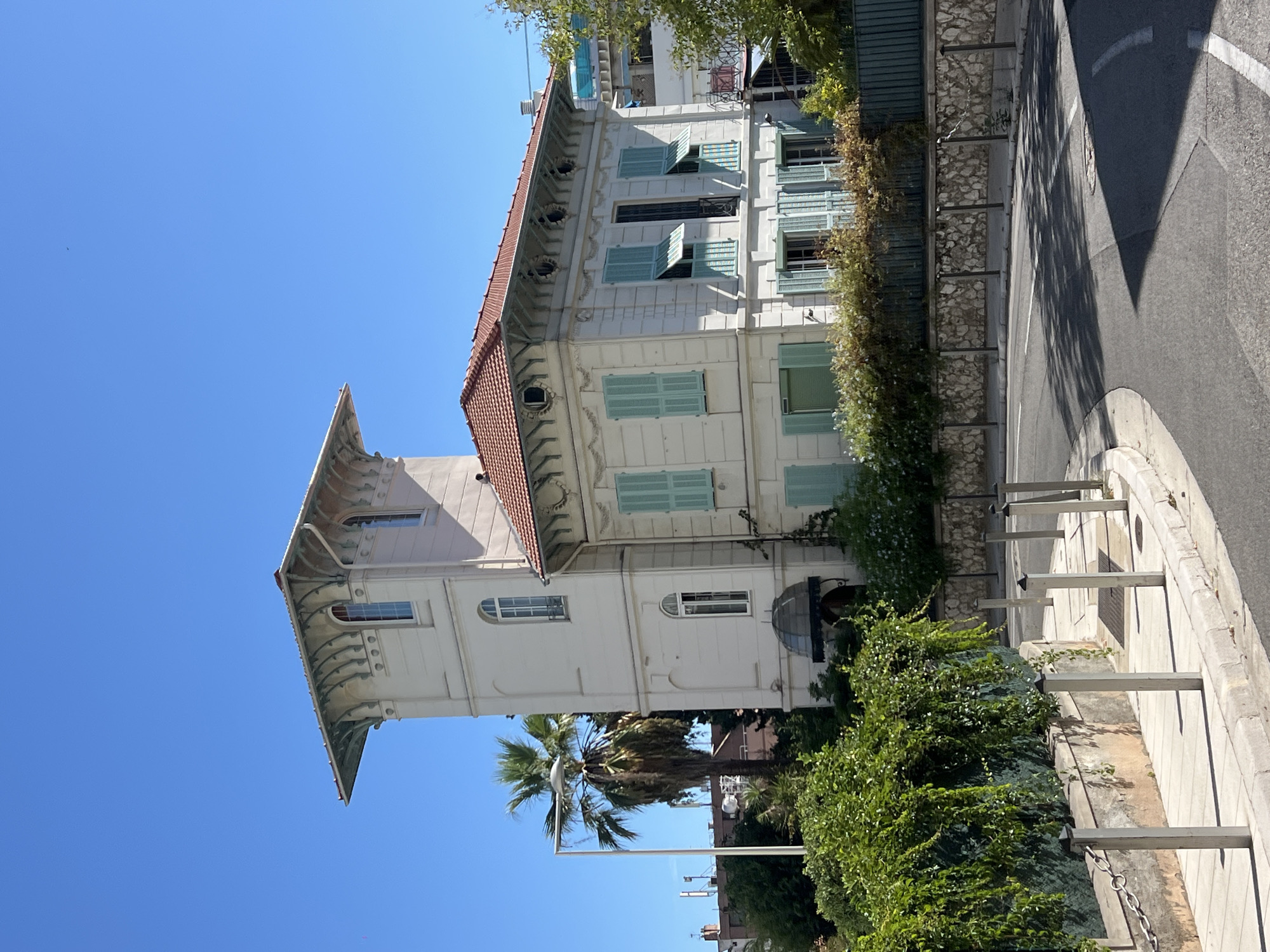 The image shows a white, multi-story building with a terracotta tiled roof in Nice, France. The building features light blue shutters on its windows and decorative architectural details, including arched windows and elaborate eaves. A tall, narrow tower section is attached to the main structure. Surrounding the building are lush green trees and a palm tree. A winding road with a sidewalk and metal bollards is visible in the foreground. The sky is a clear, bright blue.