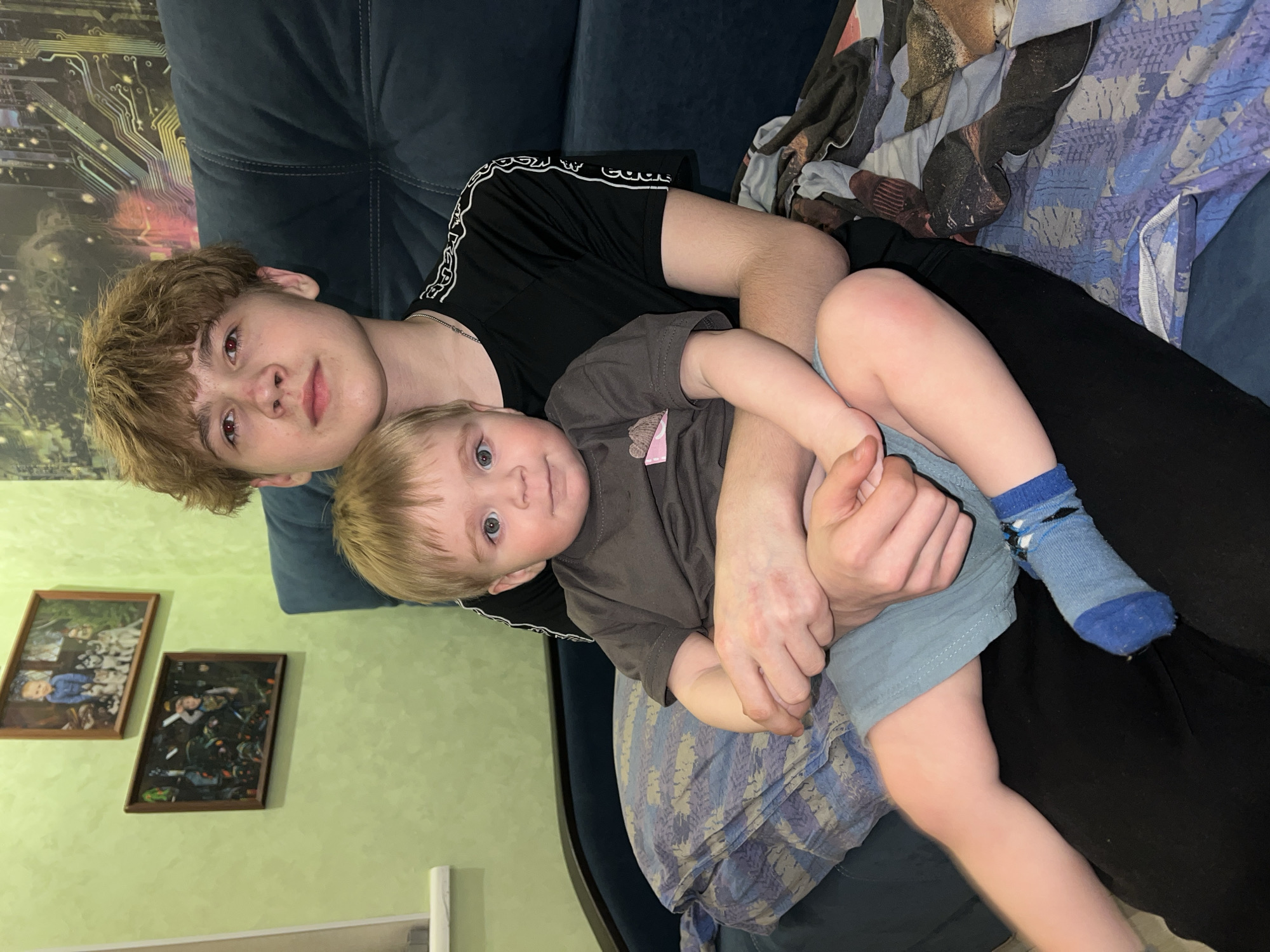 A Caucasian male with reddish-brown curly hair, fair skin, and wearing a black t-shirt with white stripes on the sleeves is lying on a dark blue couch. He is holding a toddler with fair skin and blond hair who is wearing a gray t-shirt and blue shorts with a blue sock on one foot. The toddler is looking directly at the camera. The couch is covered with a patterned blue blanket. On the wall behind the couch are two framed pictures. The wall is painted light green. The location is likely a home. It is not possible to confirm the location within Perm, Russia, from the visual information provided.