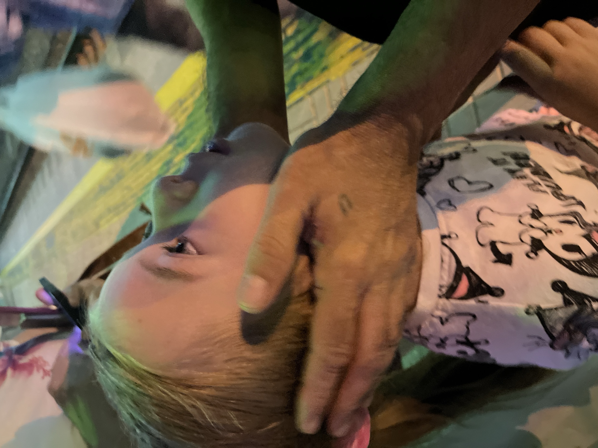 In a close-up, low-angle shot, a young girl with blonde hair pulled into pigtails looks upwards and to the right. Her face is partially obscured by two adult hands gently cupping her head and neck. The girl is wearing a light-colored t-shirt adorned with a black and pink cartoonish print. The background is out of focus, showing a yellow line on a patterned surface, suggesting an outdoor urban setting, possibly a street or promenade in Sochi, Russia, with hints of colorful lighting.