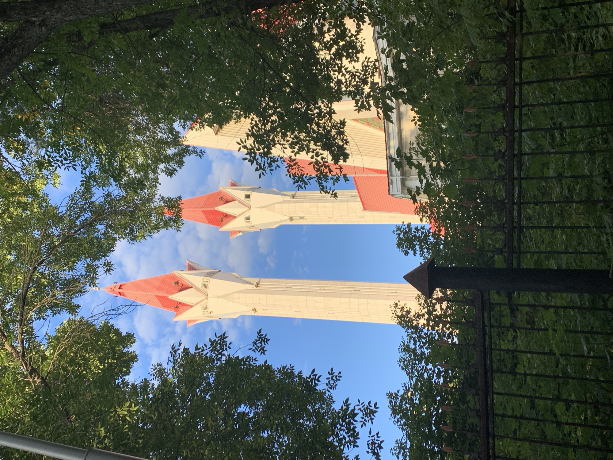 The image shows a view of the Lala-Tulpan Mosque in Ufa, Russia, from behind a dark metal fence and through lush green trees. Two tall, cream-colored minarets with pointed red roofs dominate the upper portion of the frame, silhouetted against a bright blue sky with scattered white clouds. The minarets are octagonal in shape, with decorative white detailing around their bases. A portion of the main mosque structure, also with a red roof and white walls, is visible between and behind the minarets. The foreground is filled with dense green foliage, with the tops of the trees obscuring part of the sky and the lower sections of the minarets. The fence in the foreground is made of dark metal bars with decorative finials, and a thick, dark post stands in the center of the frame, also topped with a pointed element. The scene appears to be a sunny day, with shadows cast by the trees suggesting it is either morning or late afternoon.