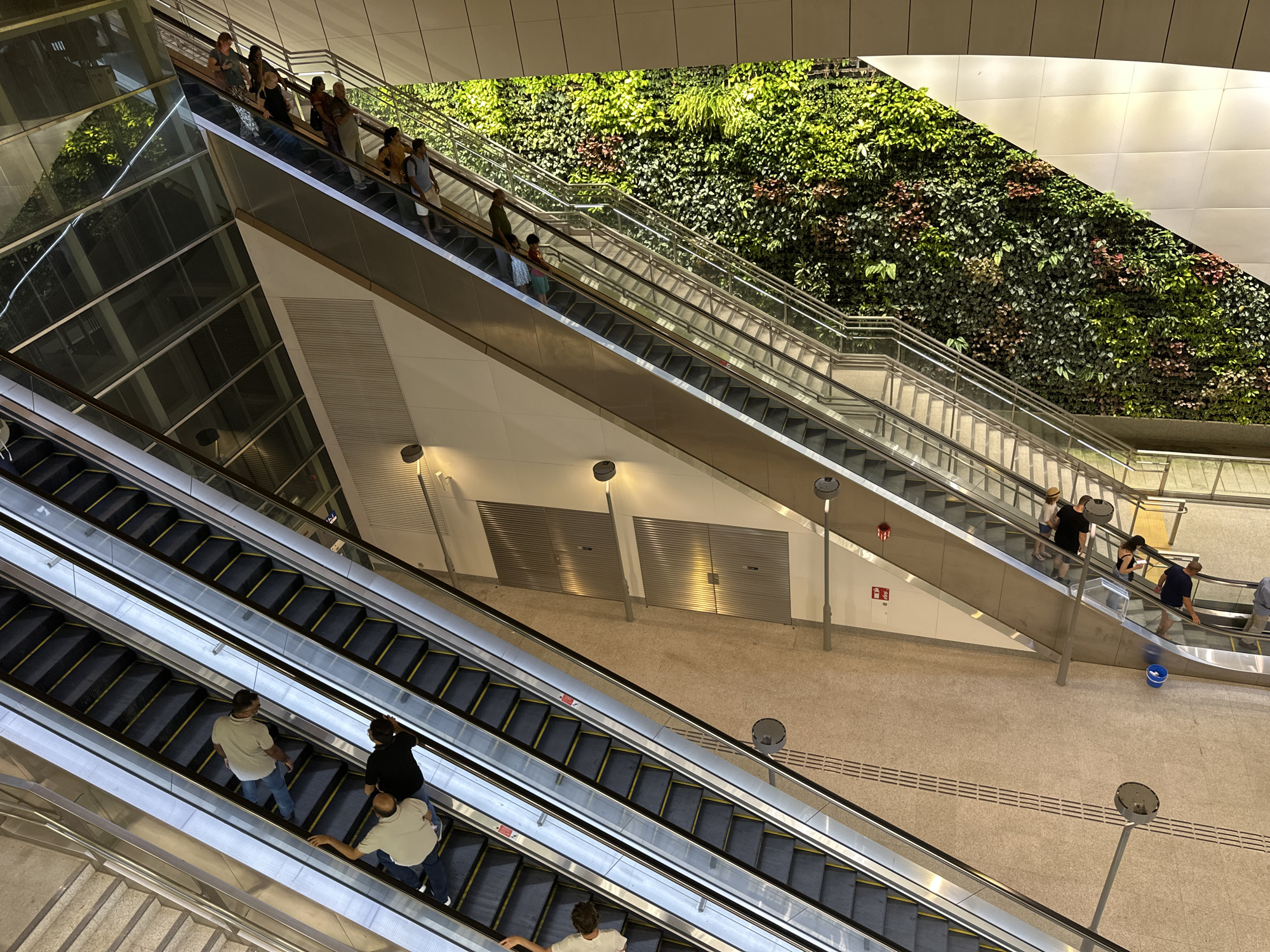 The image shows an interior scene with multiple escalators and a living wall. People are seen using the escalators, moving both up and down. The environment appears to be a modern transportation hub or public building, characterized by sleek metallic finishes, glass railings, and uniform lighting. A prominent feature is a large, lush green wall covered in various plants, adding a natural element to the artificial surroundings. The floor is tiled with a light-colored material, and several lampposts are visible, providing illumination. The overall impression is one of a clean, well-maintained, and architecturally interesting space, likely in a contemporary urban setting within Singapore.