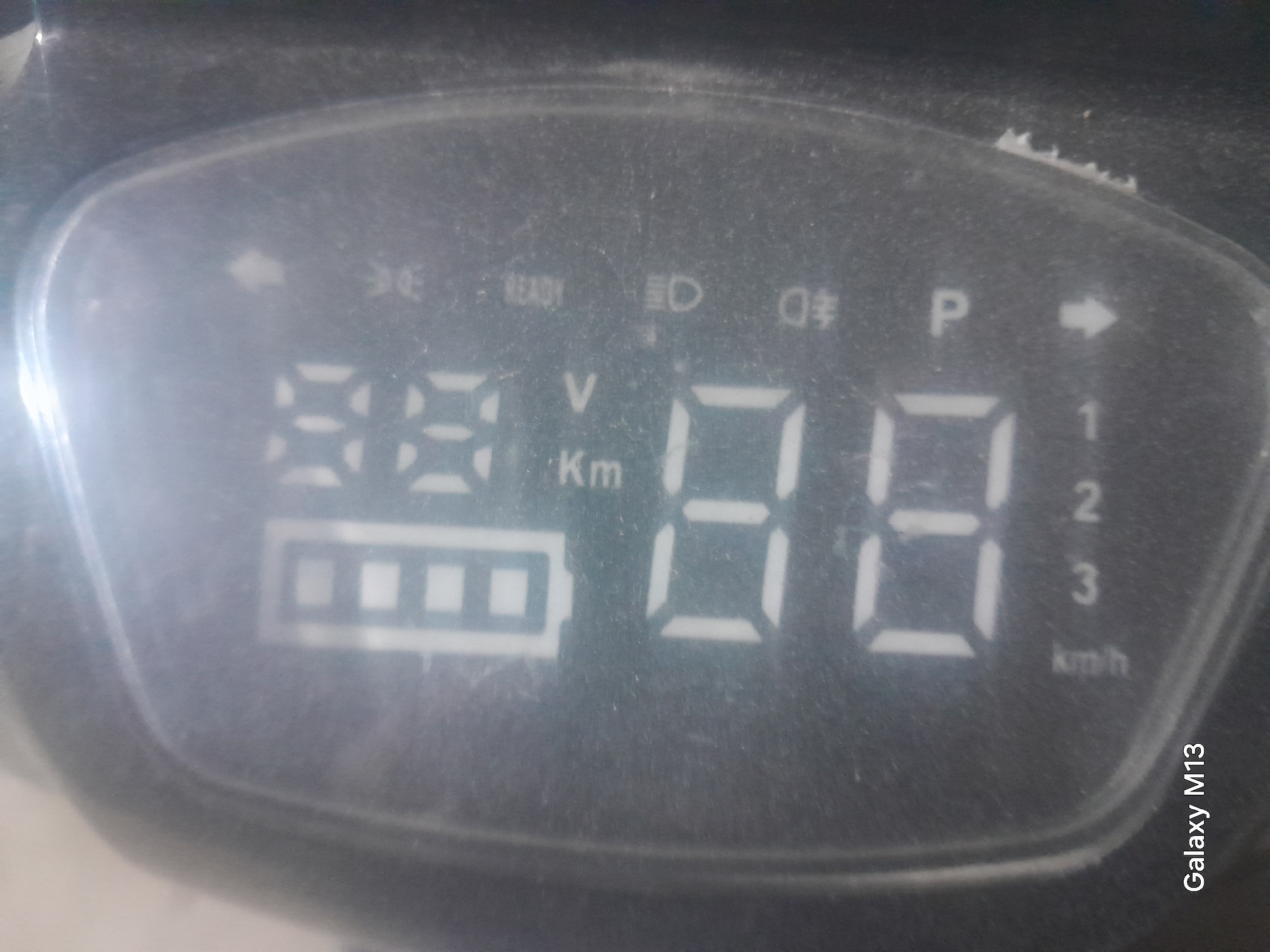 The image displays a close-up of an electronic display, likely from an electric scooter or similar vehicle. The display shows a battery indicator with several segments illuminated, suggesting a charge level. Below the battery indicator, there are large digital numbers, appearing to be "88". To the right of the numbers, there are various icons and text labels, including "Km", "V", "READY", a headlight symbol, and "P". A downward-pointing arrow is also visible. The display is smudged and dusty, indicating it is not pristine. The text "Galaxy M13" is overlaid in the bottom left corner, identifying the device used to capture the image. The overall scene is dark and lacks contextual details of a specific location in Dzharteppa, Tajikistan, as there are no discernible environmental or architectural features to suggest this location.