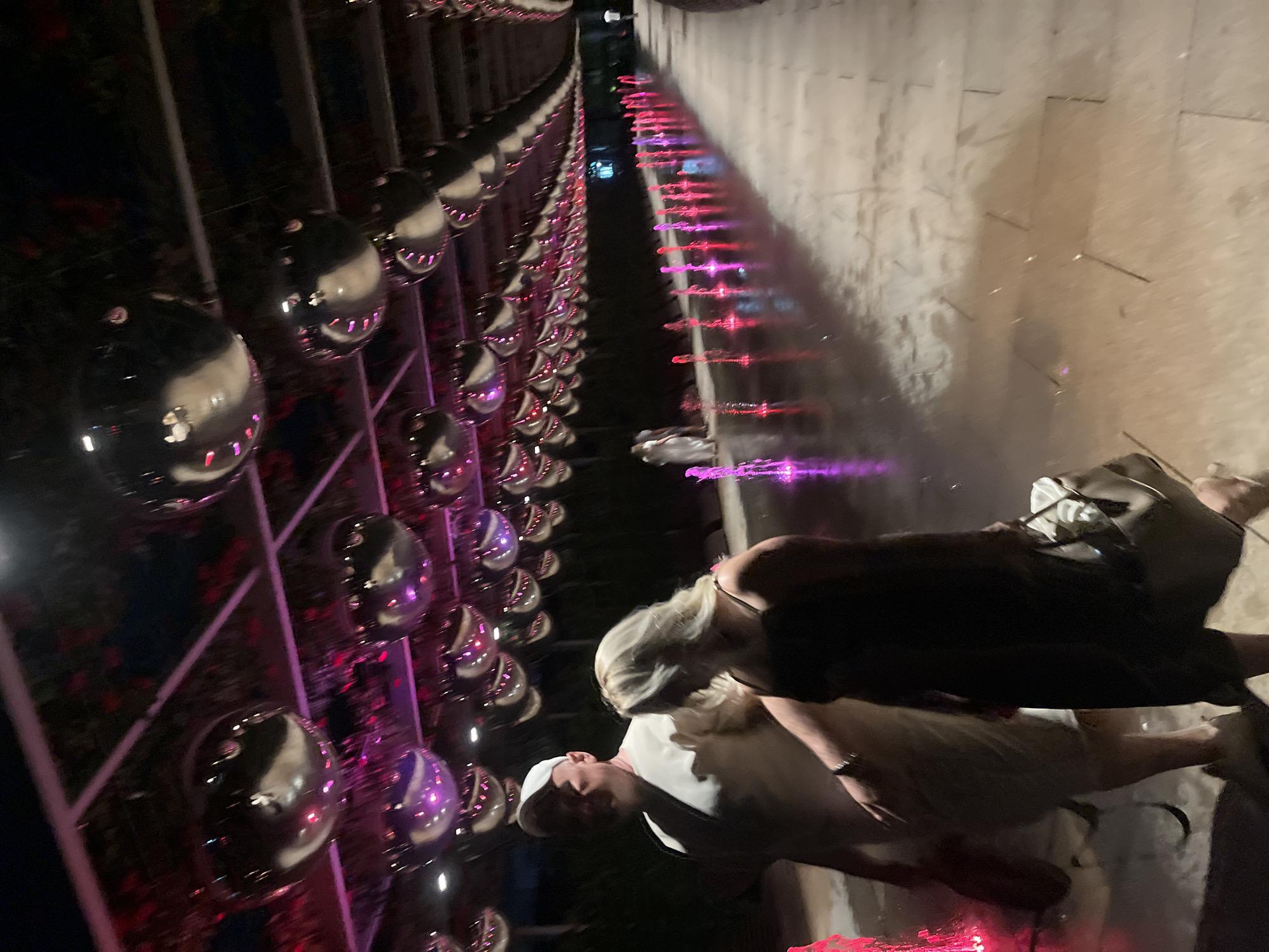 The image captures a nighttime scene at a public space featuring a water fountain exhibit. A pathway, lined with illuminated water jets in shades of pink and purple, stretches into the distance. Above, a canopy is adorned with numerous reflective silver spheres, casting a shimmering light. In the foreground, a man wearing a white cap and a light-colored outfit is walking alongside a woman with blonde hair wearing a dark dress. The woman is carrying a large handbag. Further down the pathway, other individuals are visible, including a couple dressed in white, and another person in the distance. The ground appears wet, reflecting the colored lights of the fountains. The environment suggests an outdoor public area, possibly a park or a promenade, decorated for an evening event or attraction. The presence of decorative elements like the spheres and the fountain display indicates a designed urban landscape intended for leisure or entertainment.