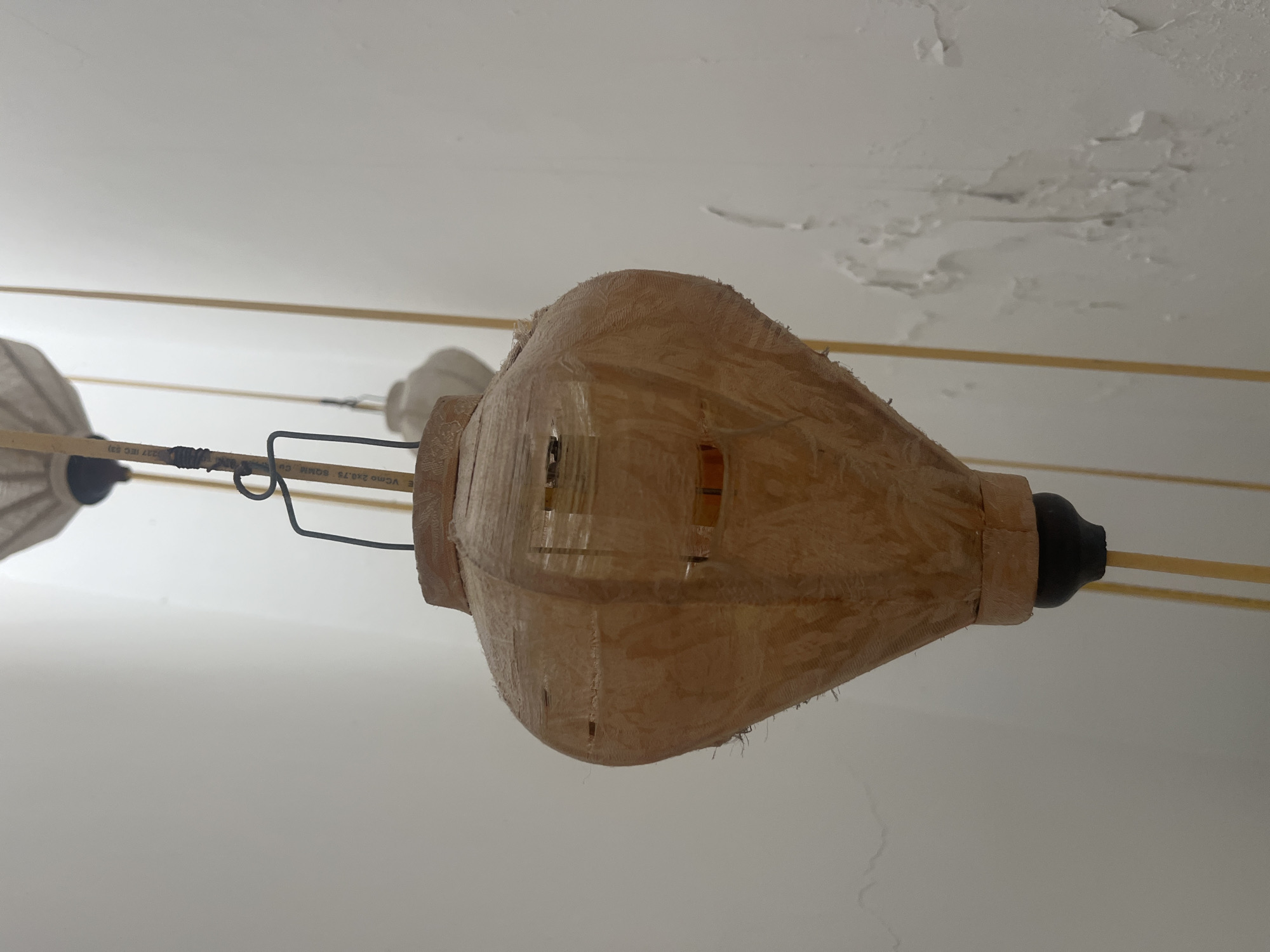 A light fixture, likely handmade, made of a woven straw-like material hangs from a wire against a light-colored wall. The light fixture is bulbous at the top and tapers to a point at the bottom, with a black cap. The material of the fixture is translucent, allowing a glimpse of what appears to be internal wiring and possibly a light bulb. The wall behind is rough and shows signs of peeling paint. In the background, other similar light fixtures hang, one with a white fabric shade and another with a similar woven material to the main subject. The scene is indoors, and the style of the light fixtures suggests they could be part of an art installation or a decorative display. The location is specified as Nha Trang, Vietnam, which aligns with the use of natural materials like straw in crafting decorative items.
