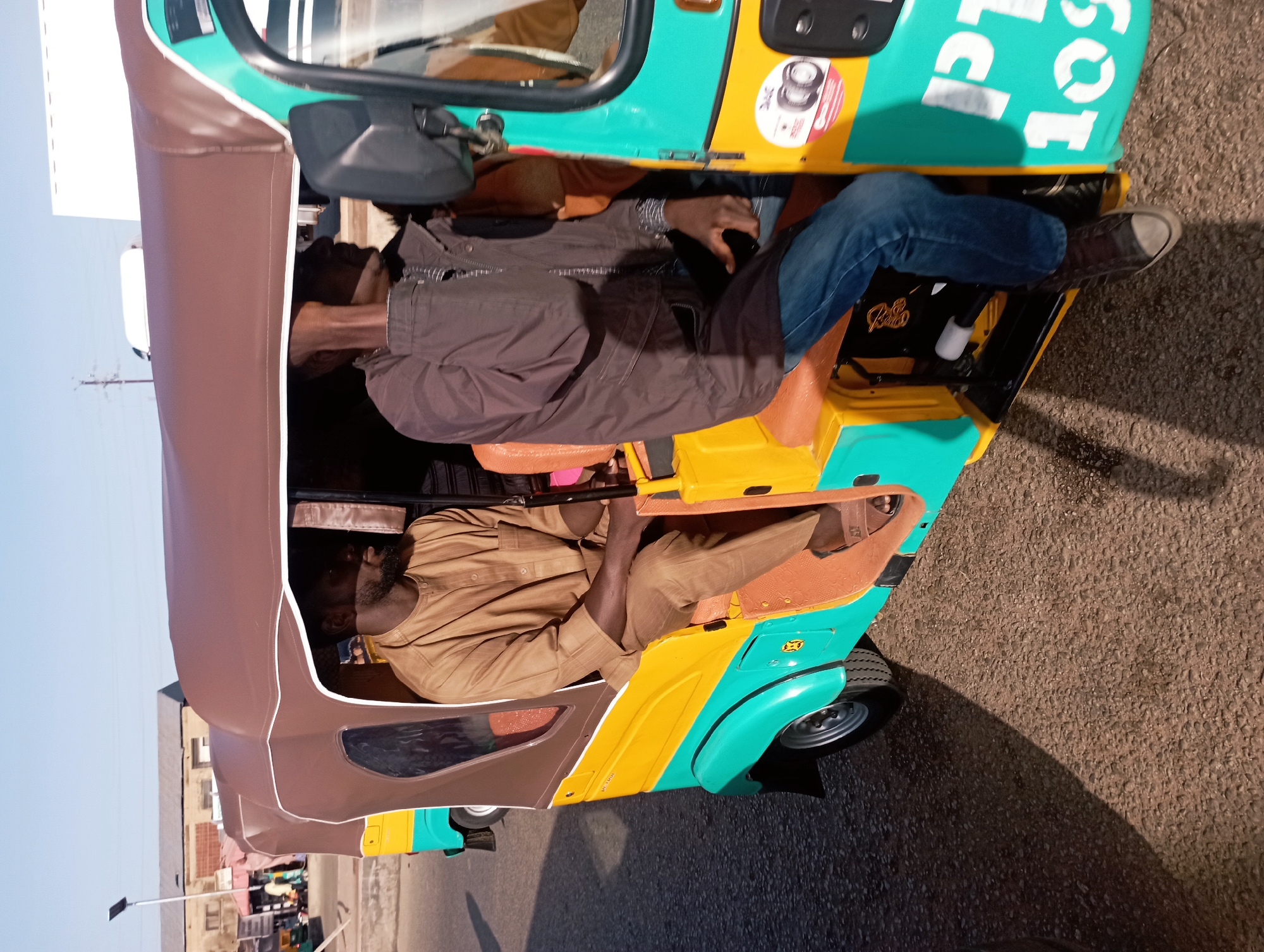 The image shows two men in a teal, yellow, and brown auto-rickshaw driving on a paved road in Jos, Nigeria. The auto-rickshaw has a brown canopy and orange seats. The man on the left is wearing a tan tunic and pants, and the man on the right is wearing a dark jacket, blue jeans, and a dark shirt with a white striped collar. Both men have beards. The man on the right is partially obscured by the frame of the auto-rickshaw. Buildings and a lamppost are visible in the background. The sun casts shadows on the road, suggesting it is daytime.