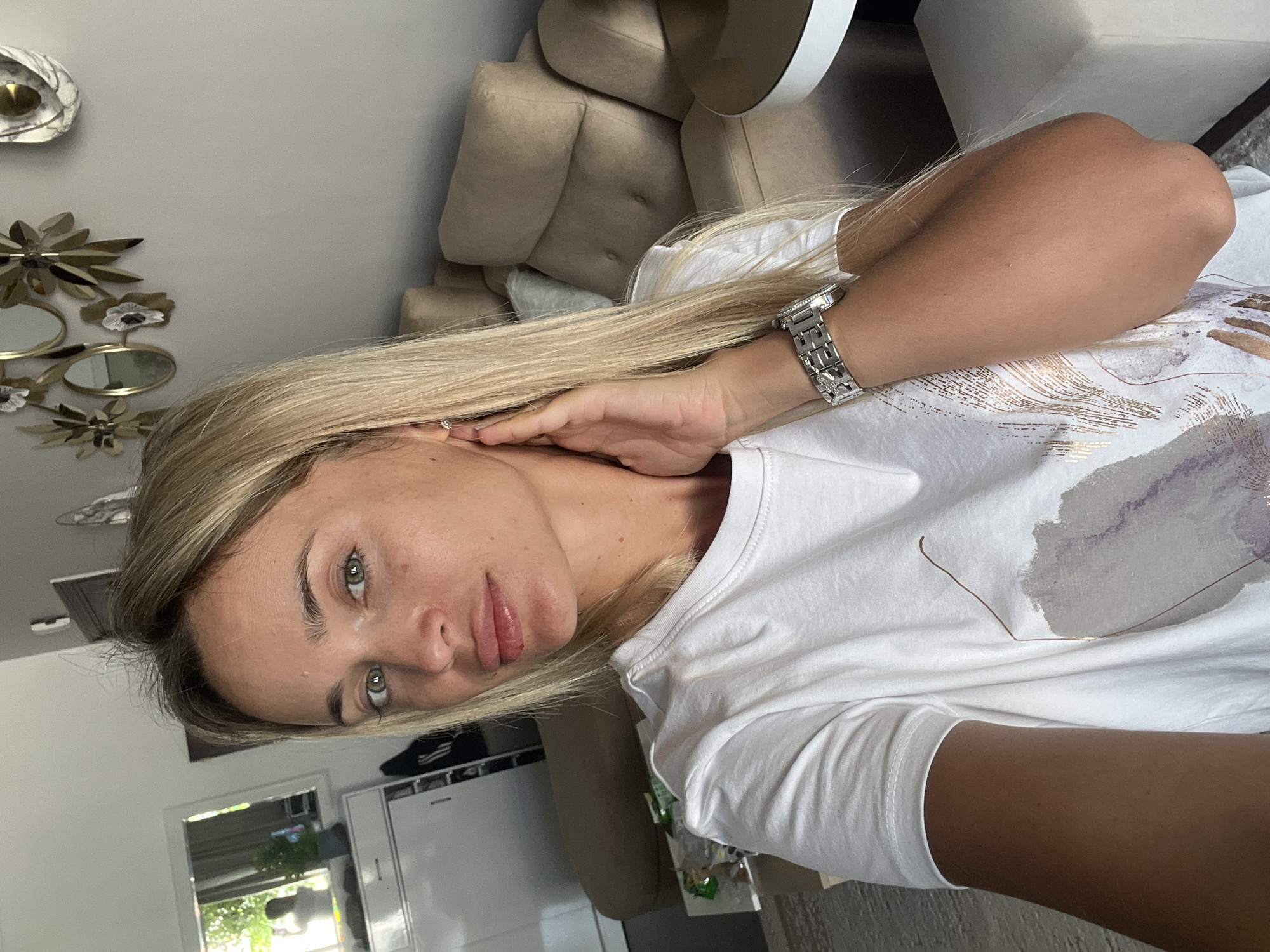 A blonde woman with green eyes is taking a selfie indoors. She is wearing a white t-shirt with a graphic design and a silver watch. Her right hand is touching her face. In the background, there is a beige sofa, a table with items on it, and a mirror reflecting a person. The environment appears to be a residential interior, possibly an apartment, though no specific location in Sharm El Sheikh, Egypt, can be definitively identified from the visual information provided.