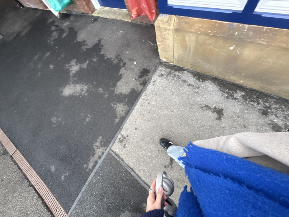From a high-angle perspective, the image captures a person standing on a paved surface. The surface is a mix of dark and light gray concrete, wet in some areas. The person is wearing jeans with frayed edges, a beige coat, and a vibrant blue scarf. They are holding a pair of gray headphones in their hand. A small section of a brick wall is visible at the top left, along with a building with blue trim at the top of the frame.