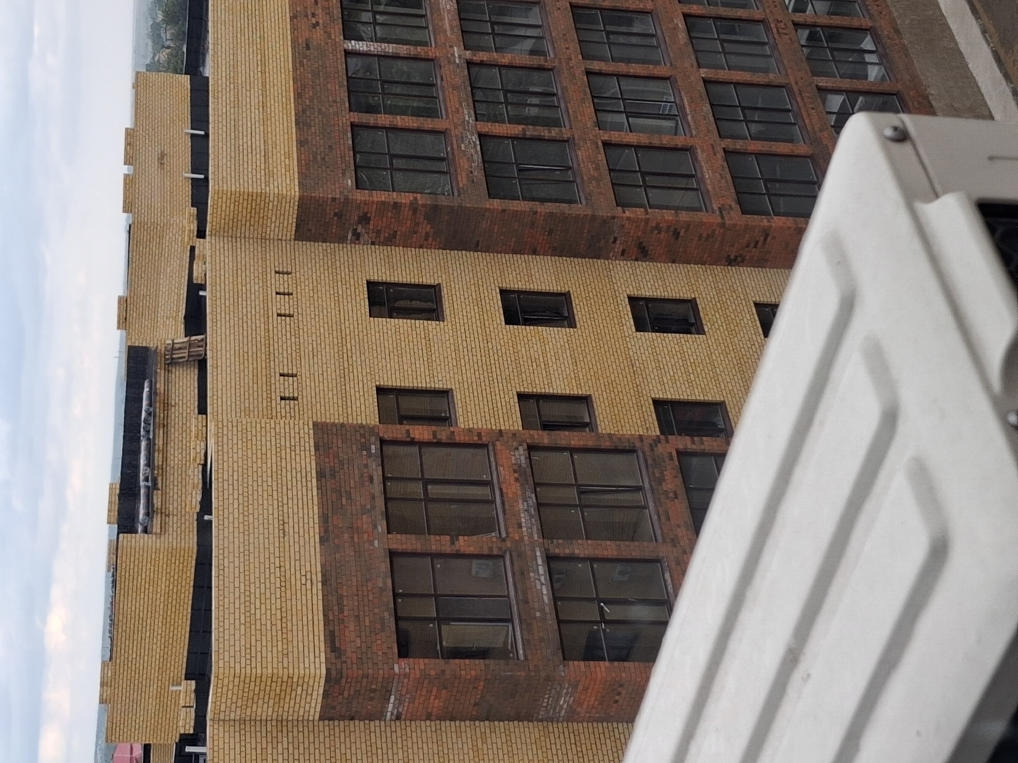 The image shows the exterior of a multi-story brick building under construction or recently completed, viewed from an elevated perspective. The building features sections of light yellow brick and sections of darker red-brown brick. Numerous windows, many appearing to be large and dark-tinted, are visible across the facades. Some windows are grouped together, forming larger openings, while others are smaller and more sporadically placed. The top of the building shows parts of the roof construction, including black roofing material and decorative brickwork along the parapet. In the foreground, a portion of a white, ribbed surface, likely part of an air conditioning unit or a similar structure, partially obscures the view of the building. The sky above is overcast with clouds. The scene is located in Semey, Kazakhstan, as indicated by the provided information. There are no people visible in the image.